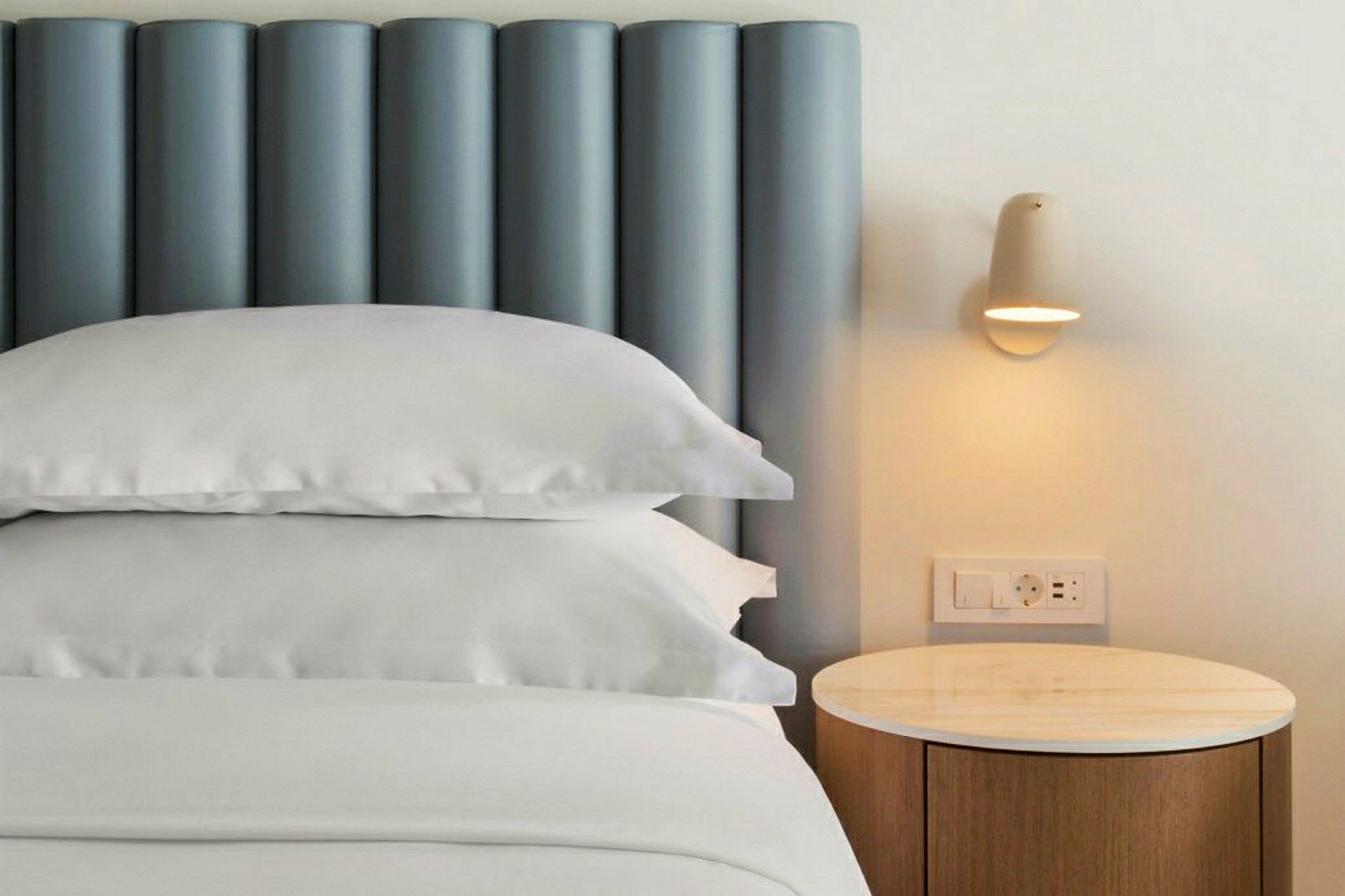 Nærbilde av en seng med blå sengegavl, hvite puter og sengetøy, ved siden av et nattbord med lampe. (AI-generert tekst)
