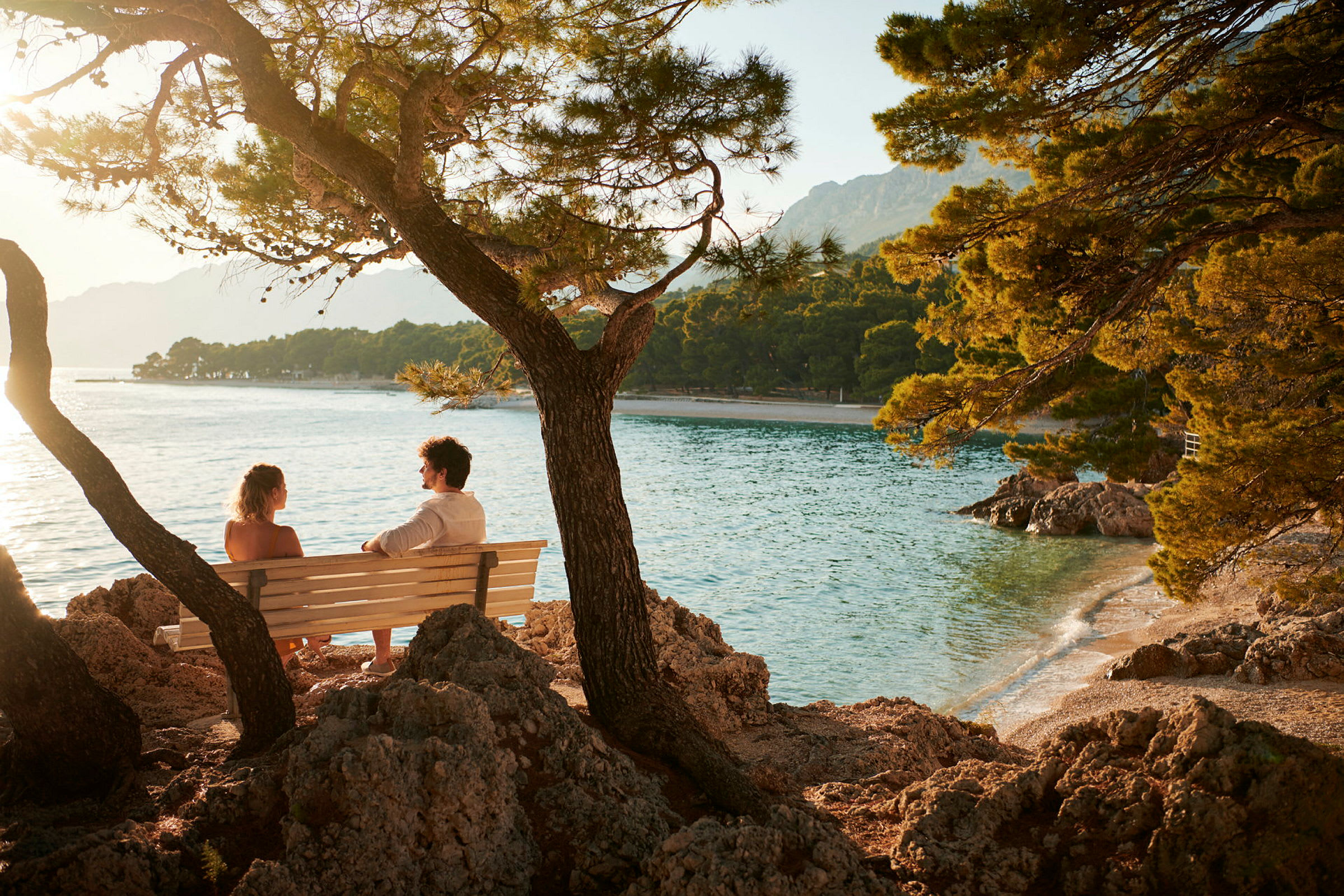 To personer sitter på en benk ved kysten med utsikt over havet og trær. (AI-generert tekst)