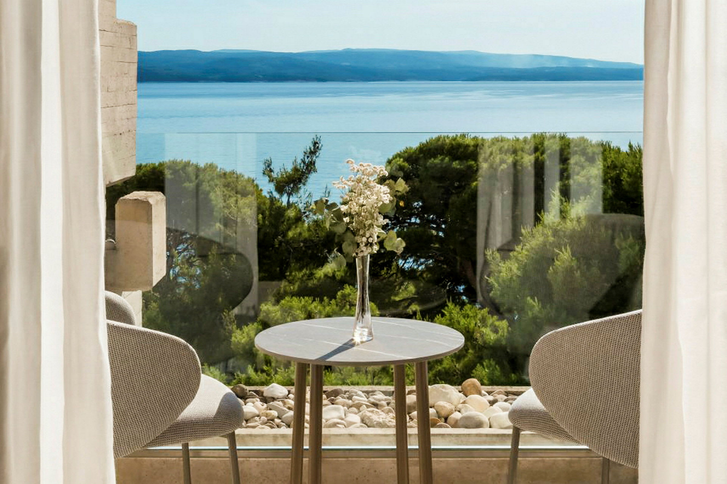 En borddekorasjon med blomster ved et bord foran vinduer med utsikt over havet og vegetasjon. (AI-generert tekst)