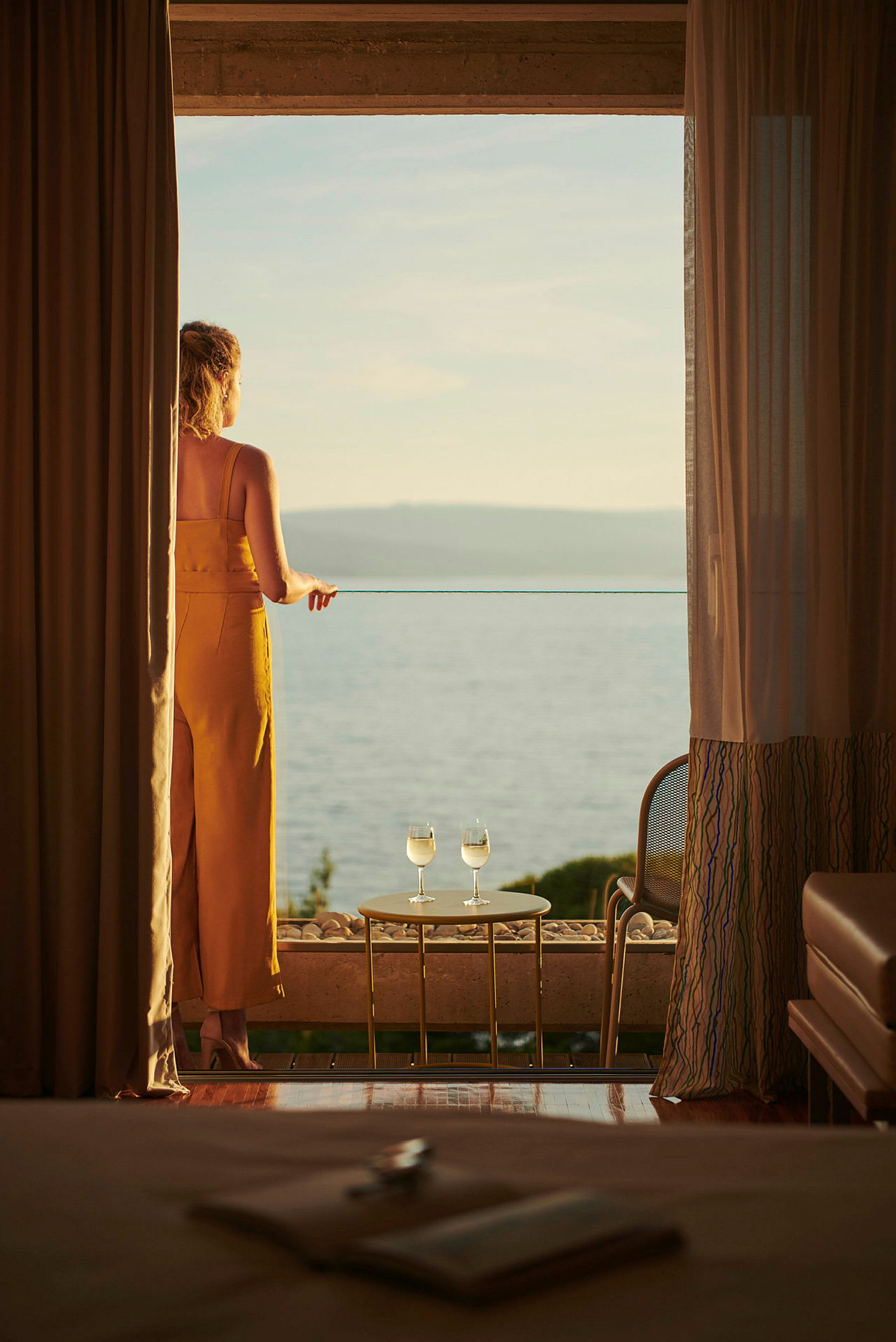 En kvinne står på en balkong og ser ut over havet, med to glass hvitvin på et bord i nærheten av henne. (AI-generert tekst)