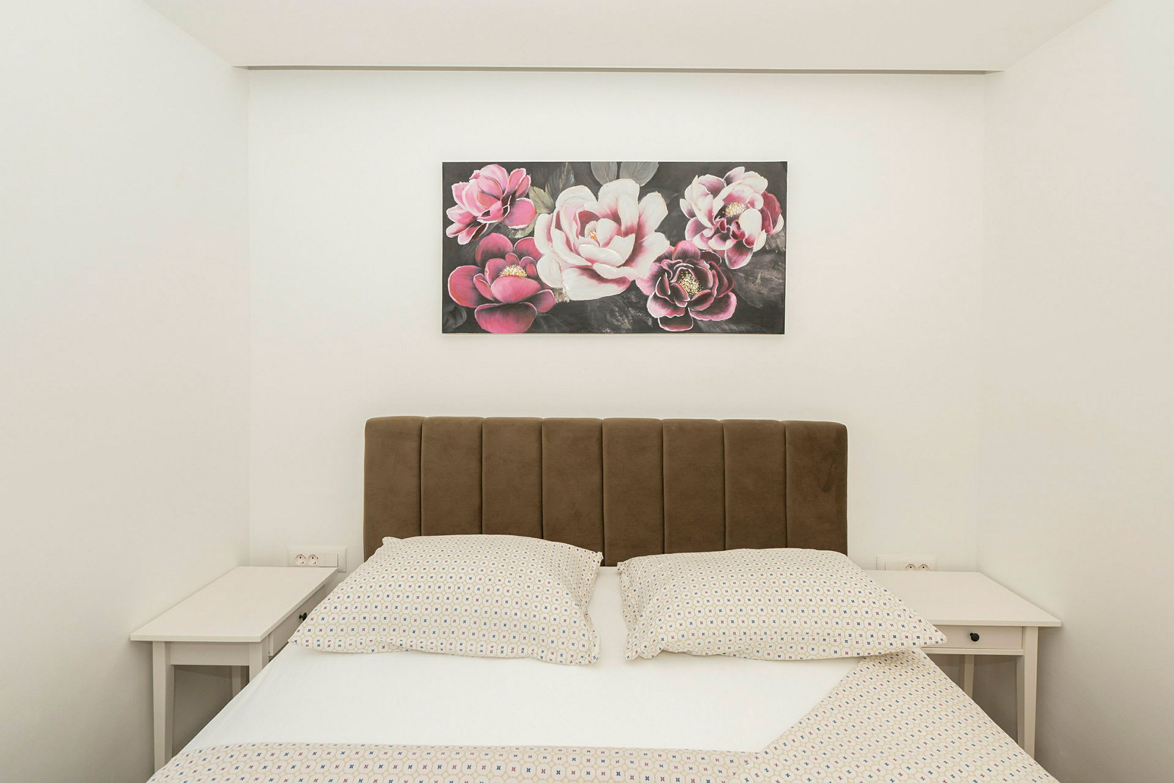 Seng med brun hodegavl, to nattbord og et maleri med blomstermotiv over. (AI-generert tekst)