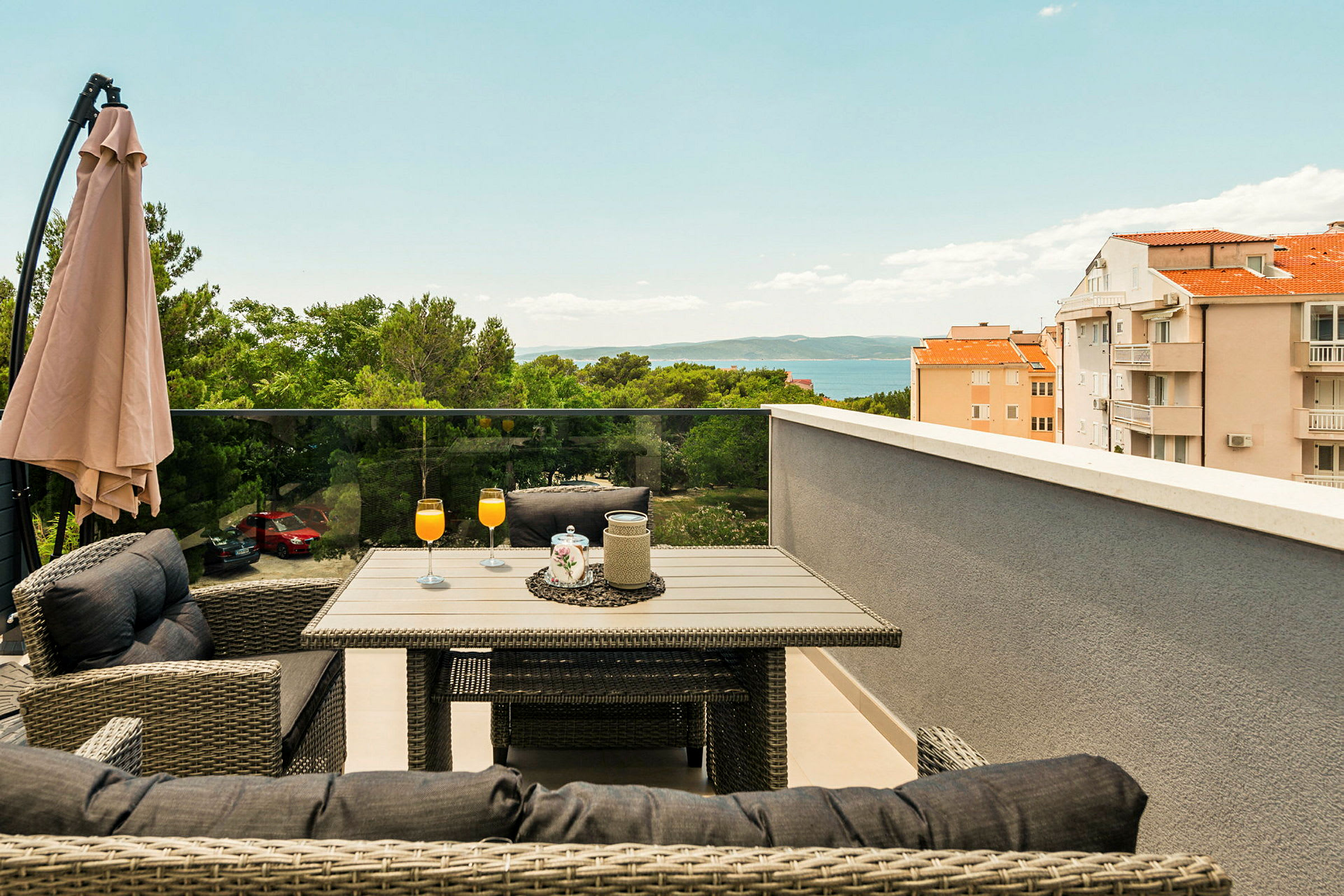Terrasse med bord og stoler, oransje drikker i glass, parasoll og utsikt over trær, bygninger og hav. (AI-generert tekst)