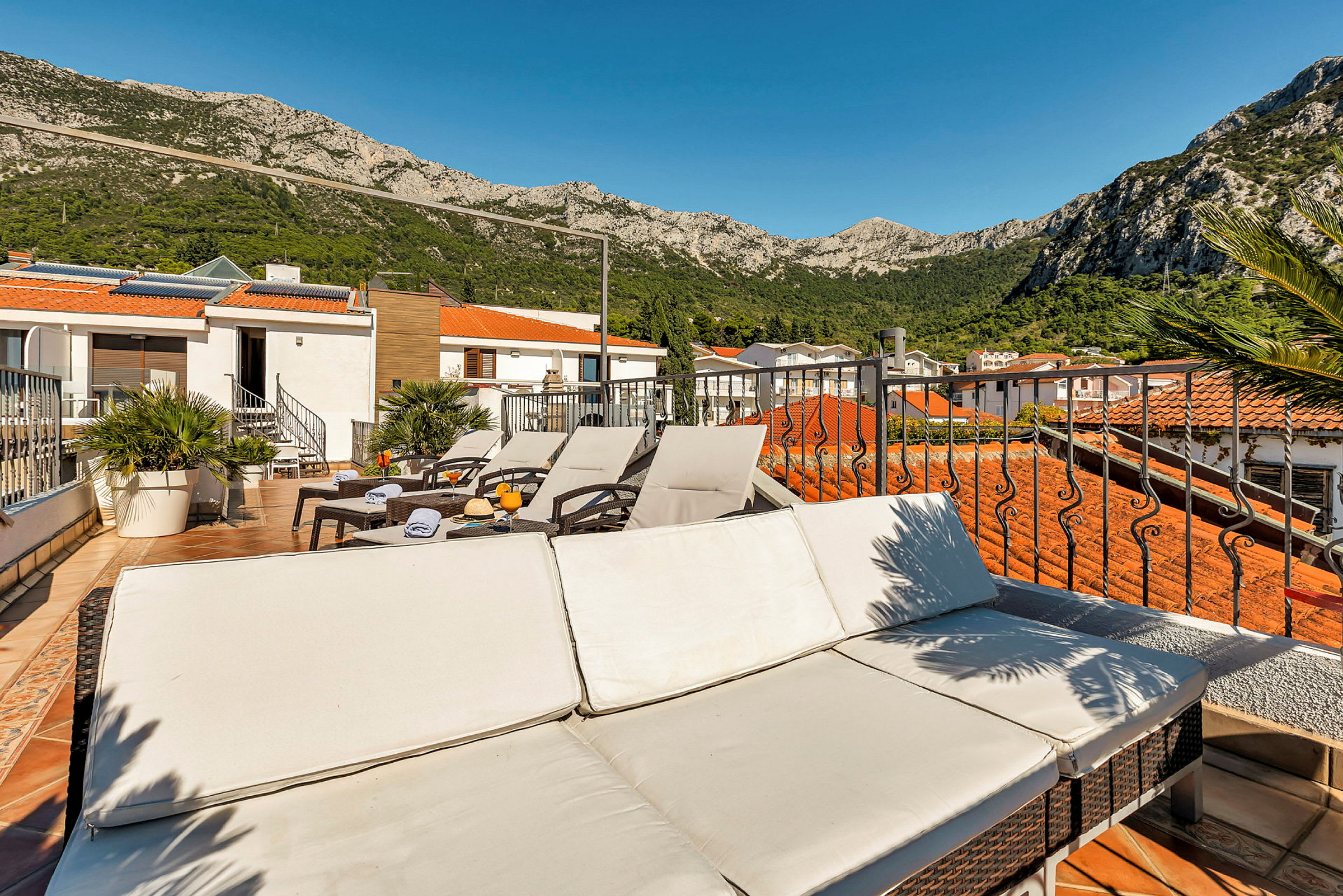 Utsikt over en terrasse med solsenger og bord, omgitt av røde tak og fjellandskap. (AI-generert tekst)