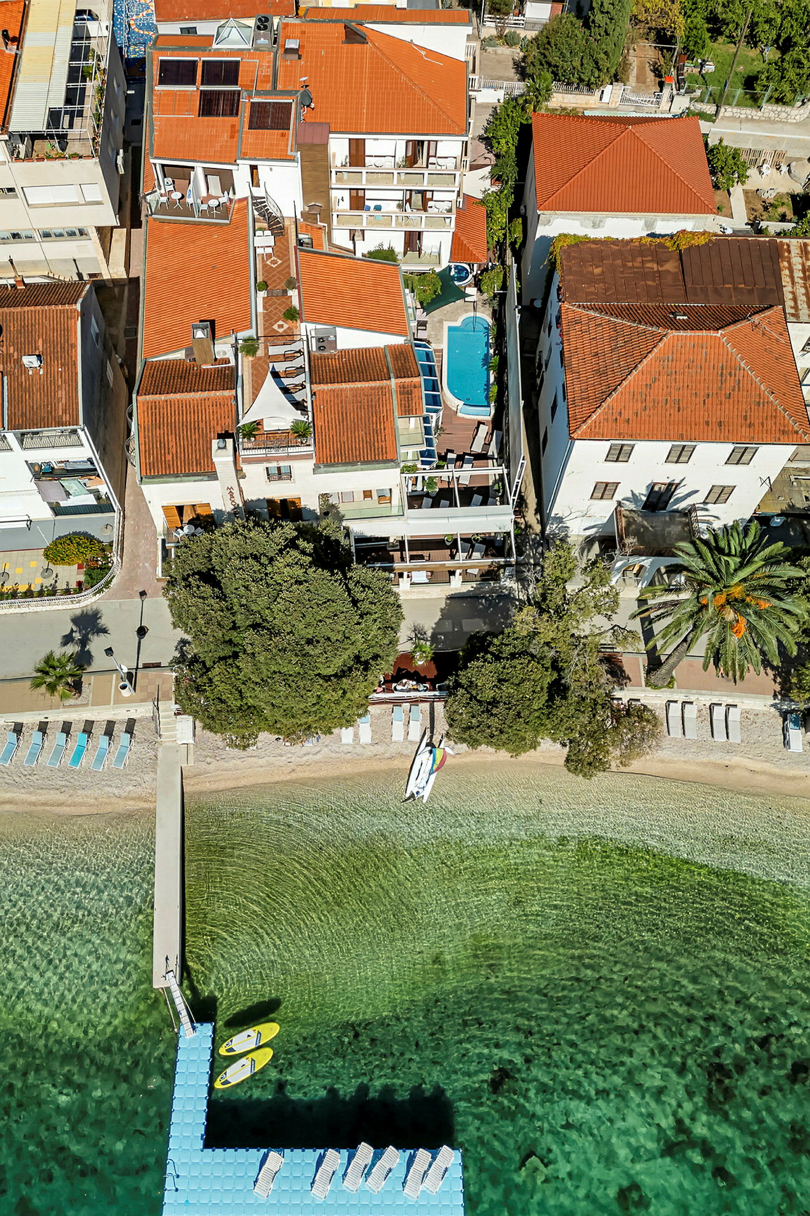 Luftfoto av en bygning med en takterrasse, et basseng og en brygge med kajakker ved en strand. (AI-generert tekst)