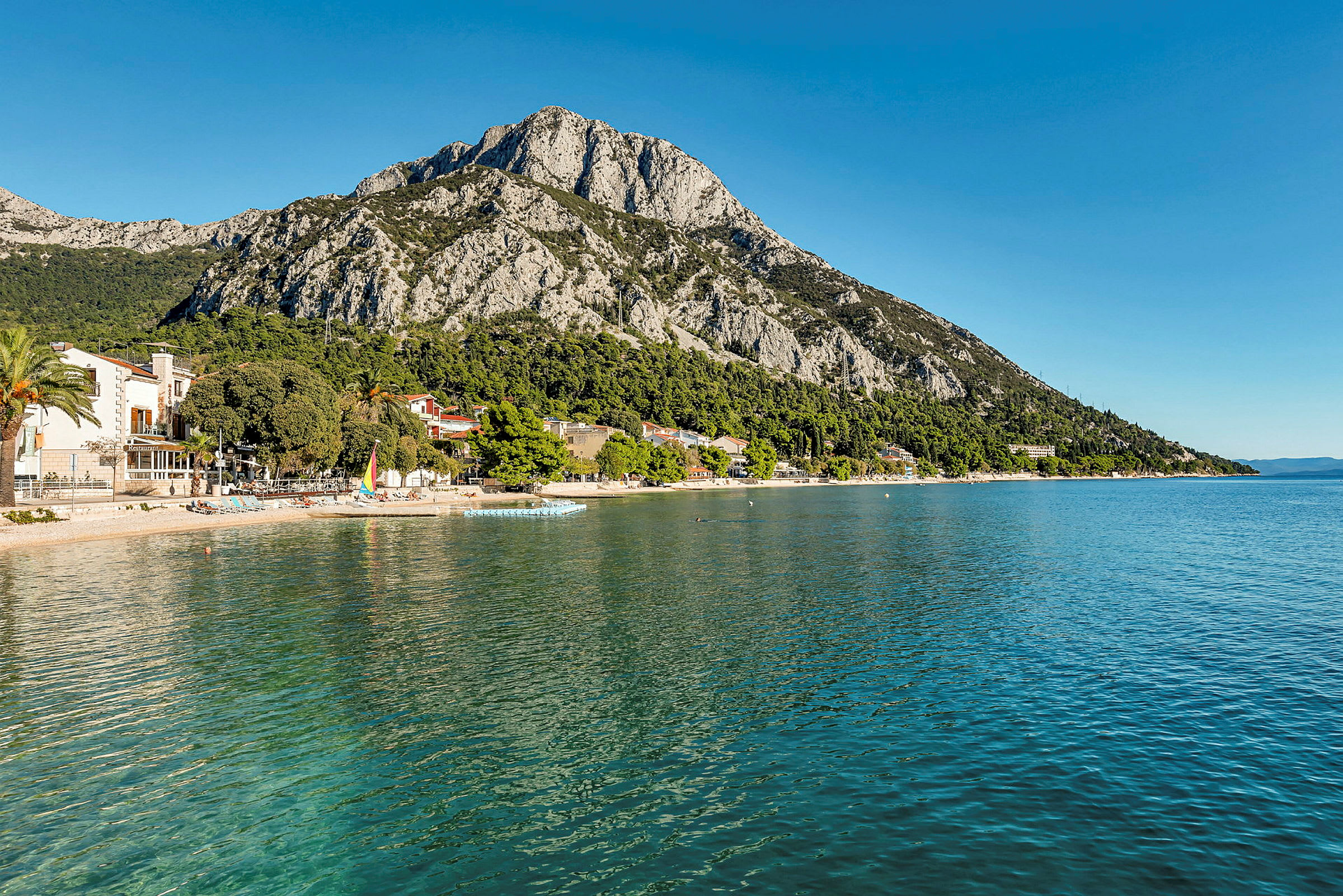 Strand med små hus ved foten av et fjell, havet strekker seg fremover. (AI-generert tekst)