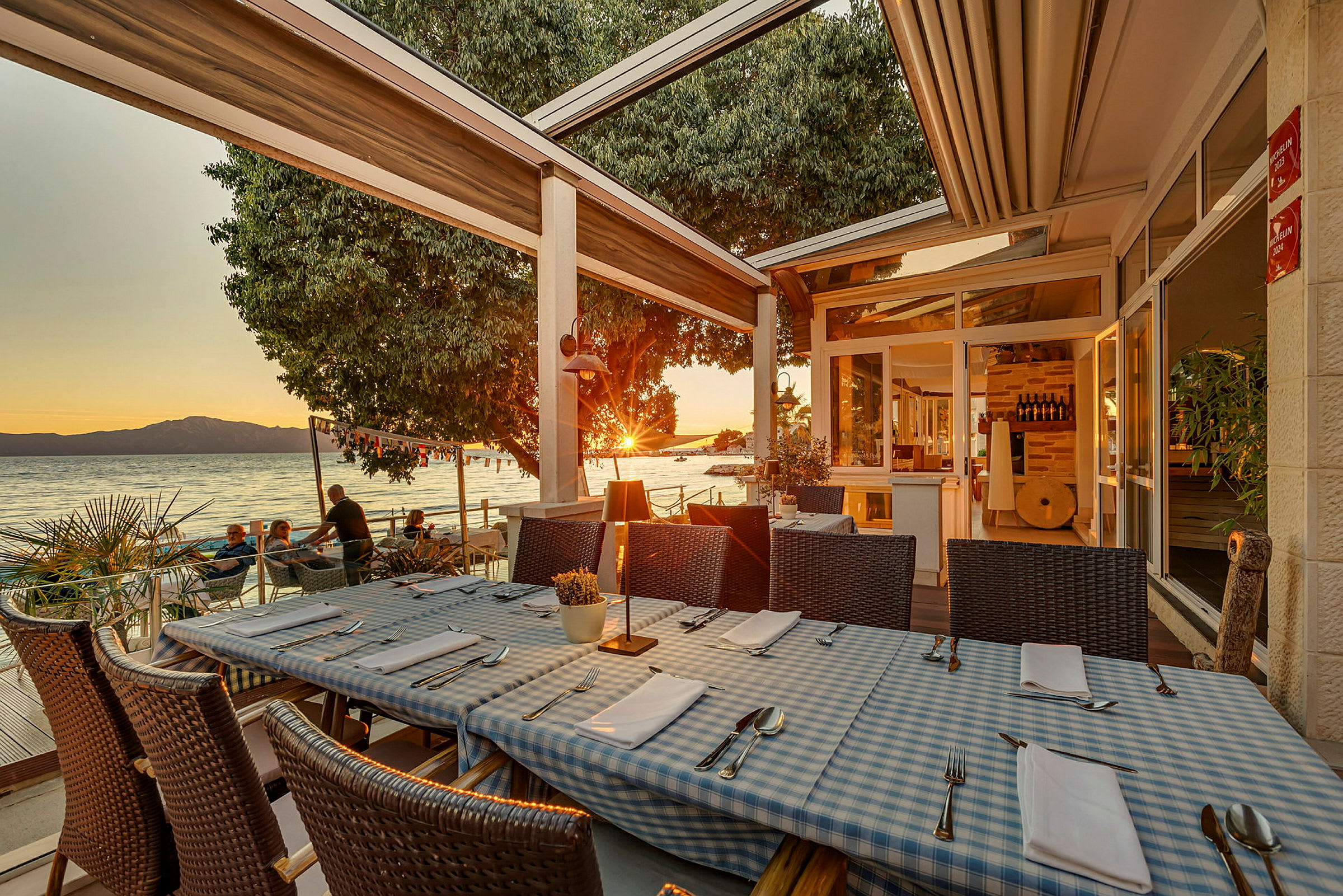 Restaurantbord med blå- og hvitrutete duker på en terrasse ved sjøen under solnedgang. (AI-generert tekst)