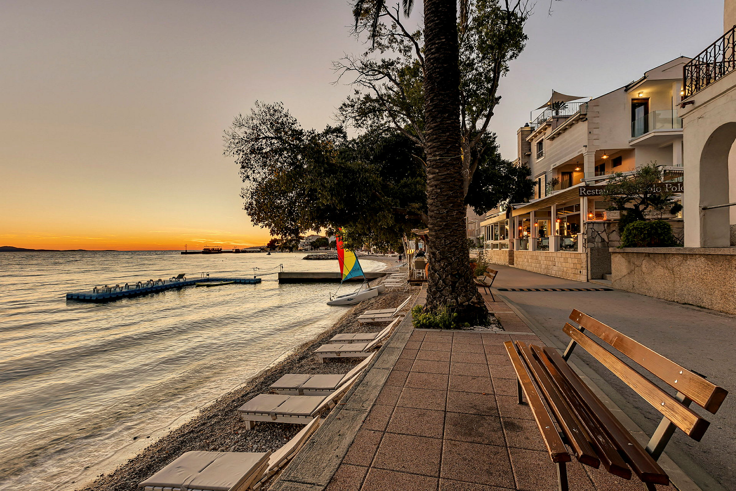 Strandpromenade med en benk, solsenger og et seil ved et rolig hav ved solnedgang. (AI-generert tekst)