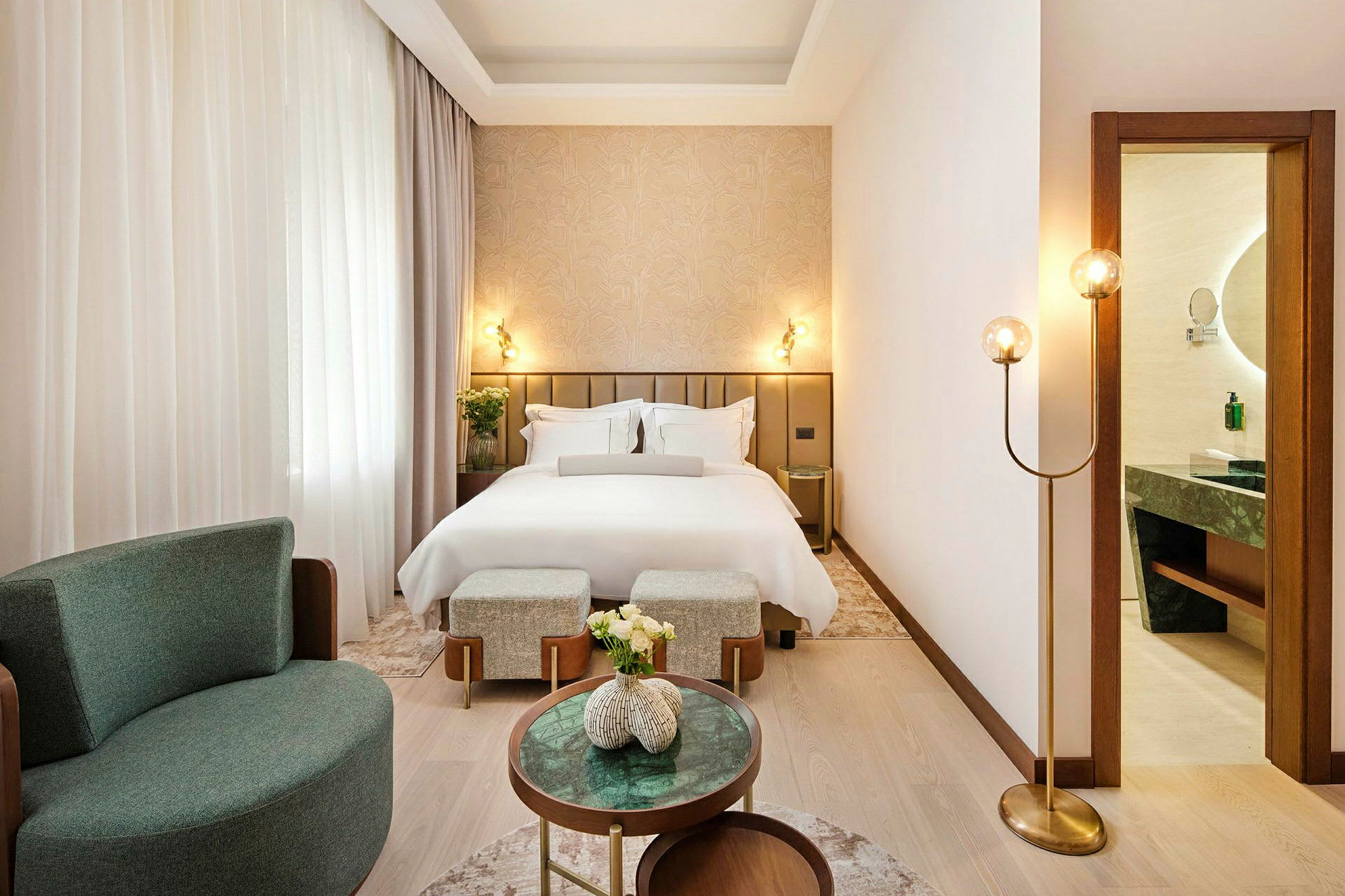Et elegant hotellrom med en oppredd seng, lenestol og små bord. Et bad ses gjennom en åpen dør. (AI-generert tekst)
