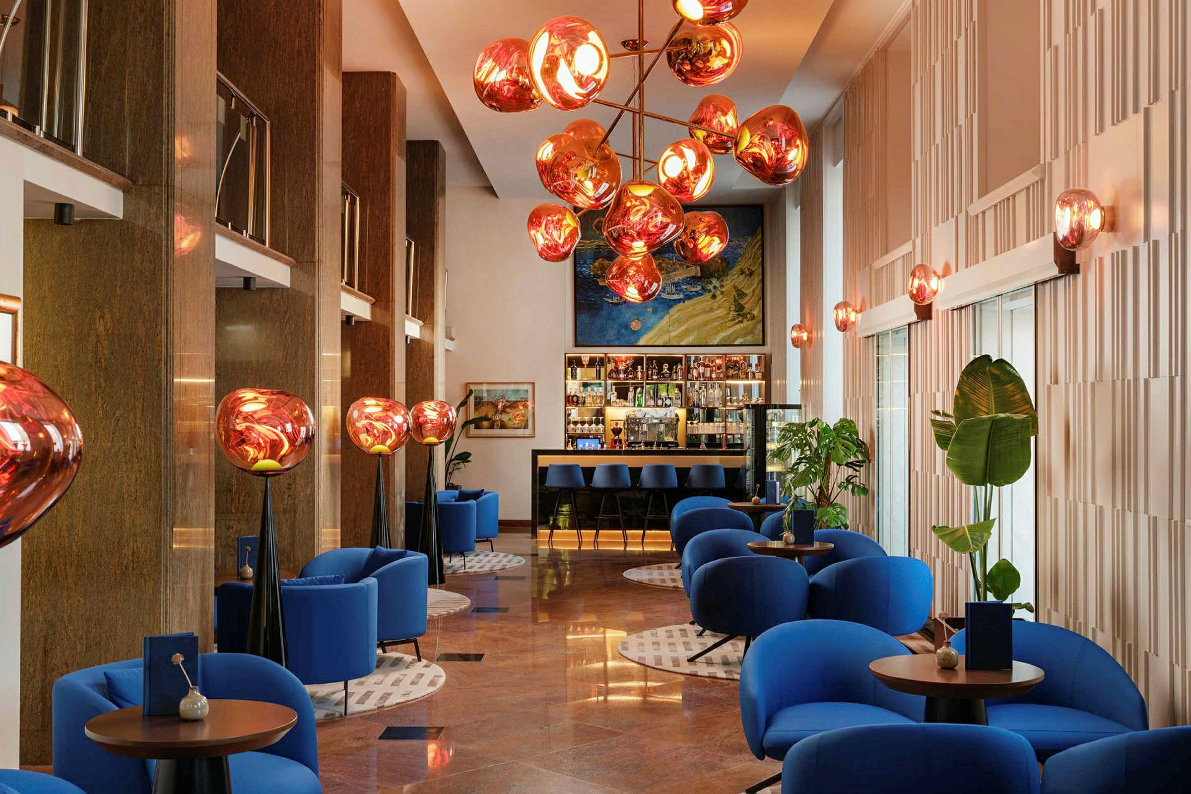 En moderne hotellobby med blå lenestoler, runde bord og dekorativ belysning i taket. (AI-generert tekst)