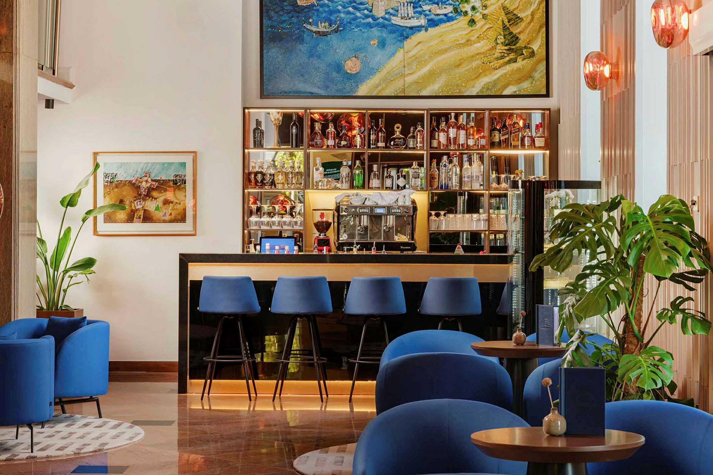 En hotellbar med blå stoler ved disken samt blå lenestoler foran små bord. Veggdekorasjoner og planter finnes i bakgrunnen. (AI-generert tekst)