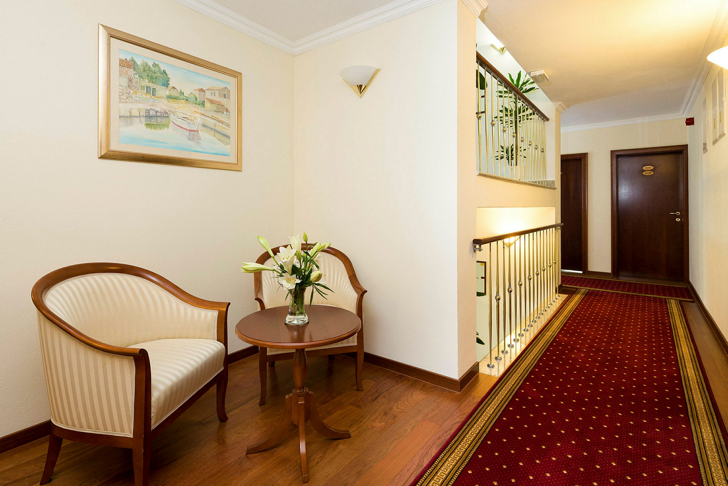 Bildet viser en hotellgang med sitteområde, inkludert to stoler, et sidebord med blomster og et rødt teppe. (AI-generert tekst)
