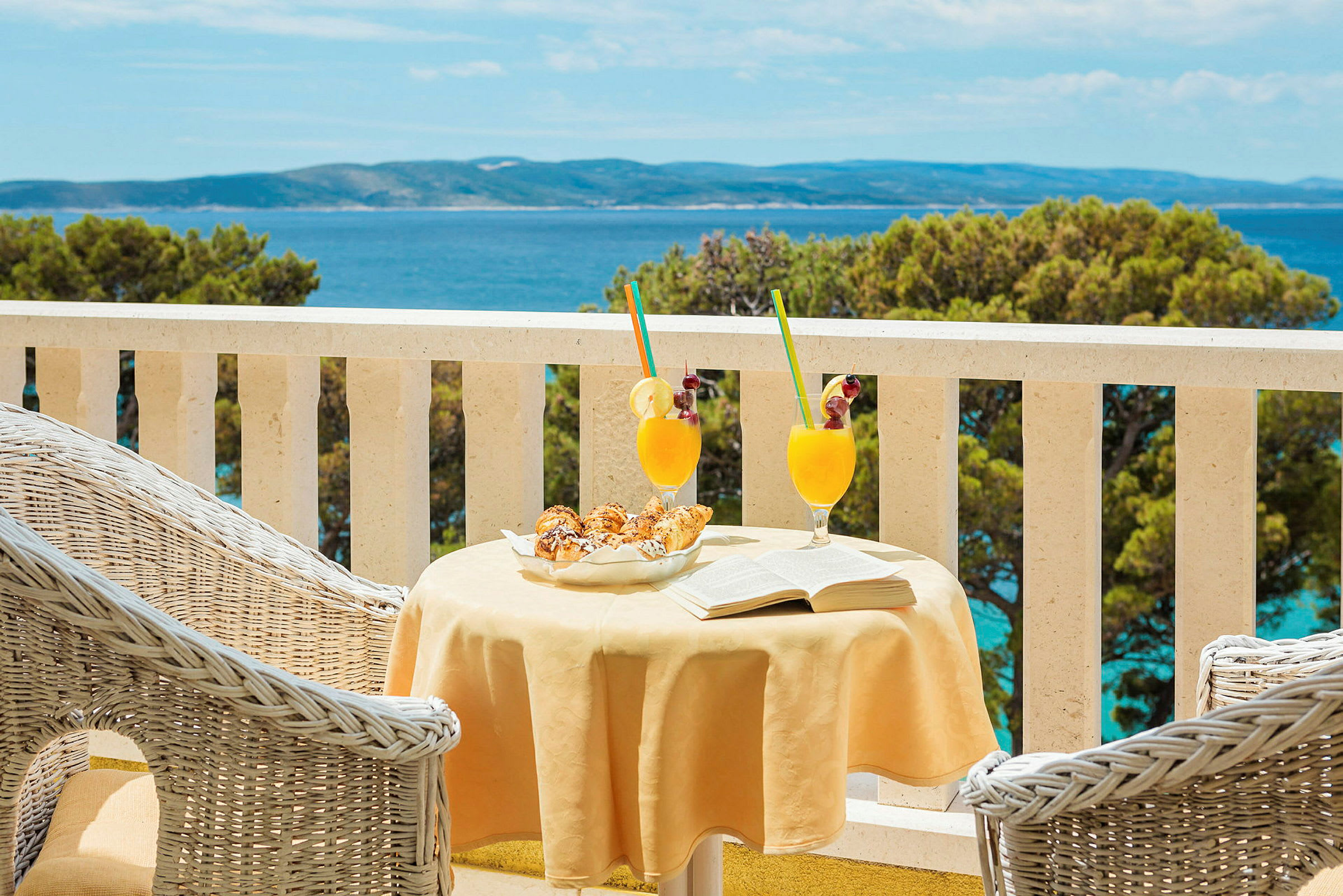 Et bord på en terrasse med to glass med oransje drikke, bakverk og en åpen bok. Utsikt over havet i bakgrunnen. (AI-generert tekst)