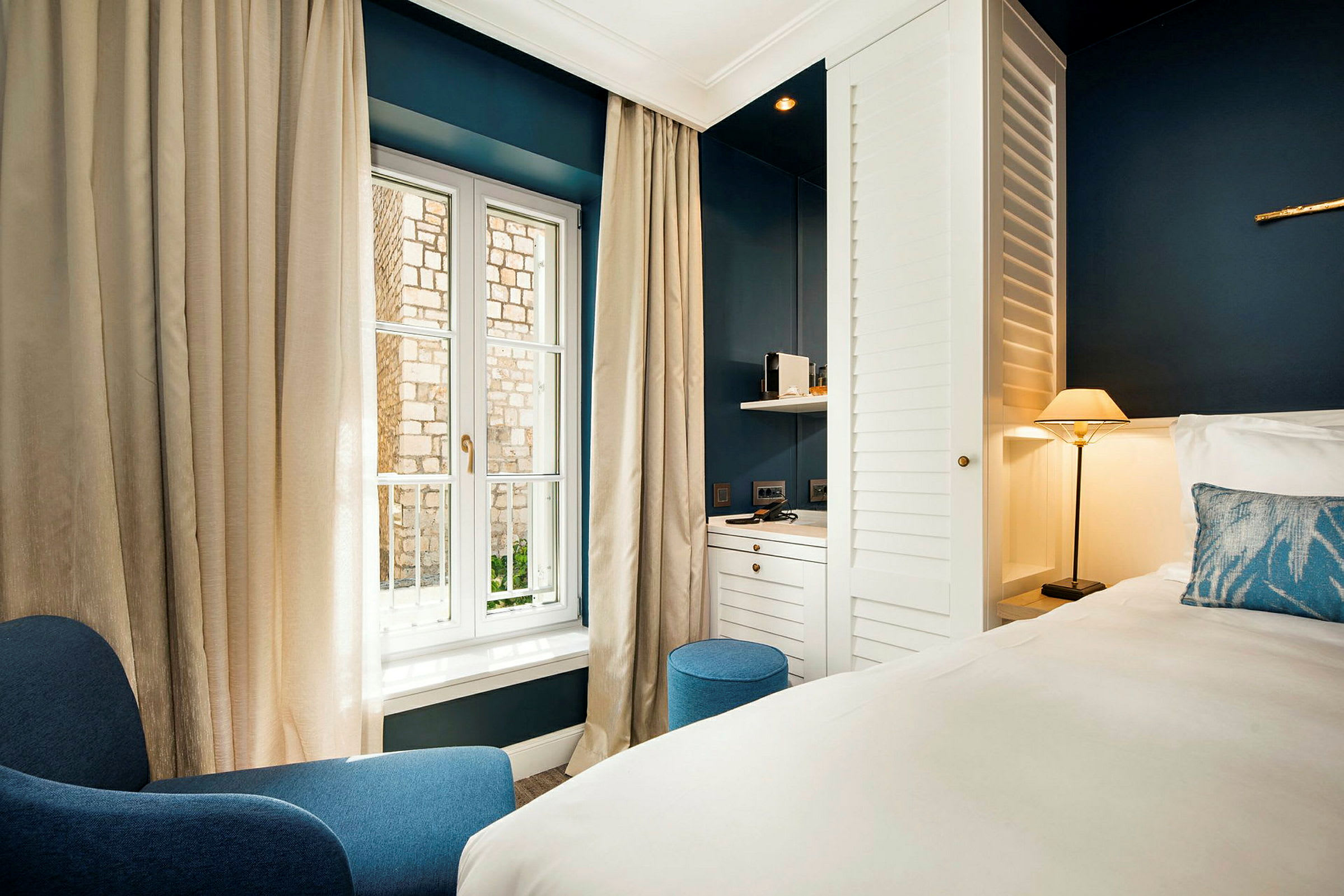 Et hotellrom med moderne møbler inkludert en blå lenestol, en seng med hvitt sengetøy, en liten lampe og et skrivebord ved et vindu. (AI-generert tekst)
