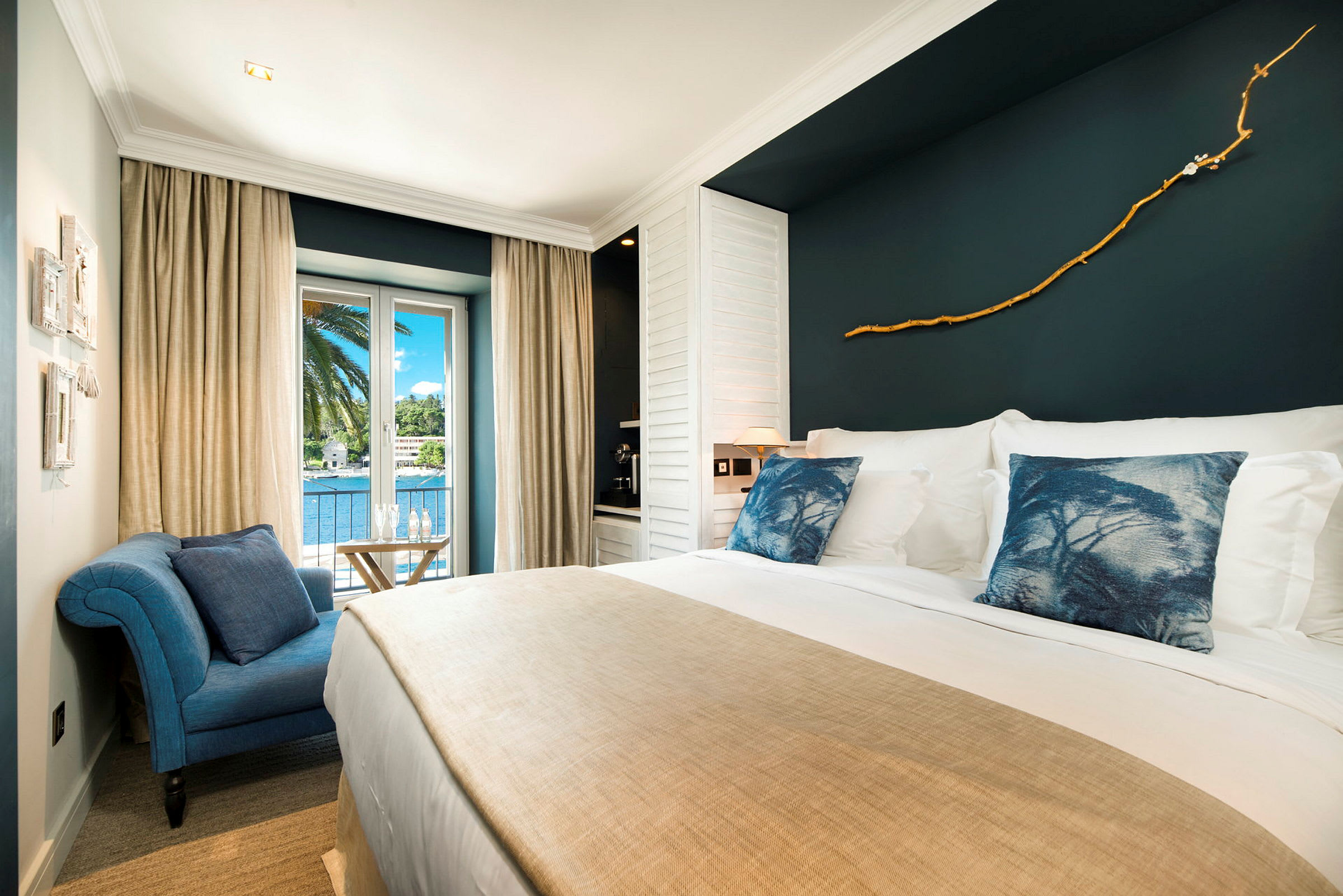 Et elegant hotellrom med stor seng, blå pynteputer, beige sengeteppe og dekorert mørk vegg. Rommet har et vindu med utsikt over palmetrær og vann. (AI-generert tekst)