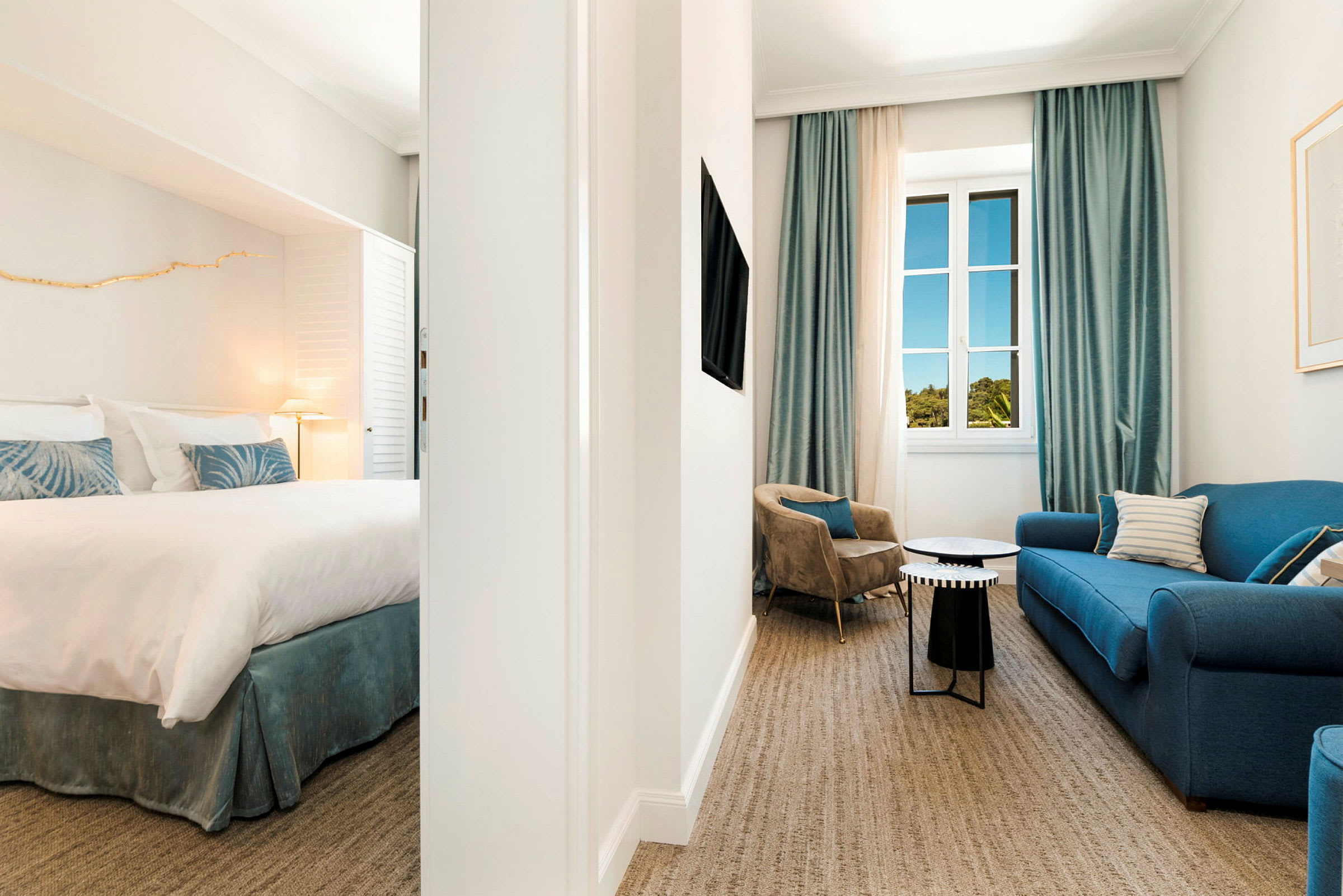 En hotellsuite med soverom og stuedel, innredet med en blå sofa, lenestoler og vindu med blå gardiner. (AI-generert tekst)