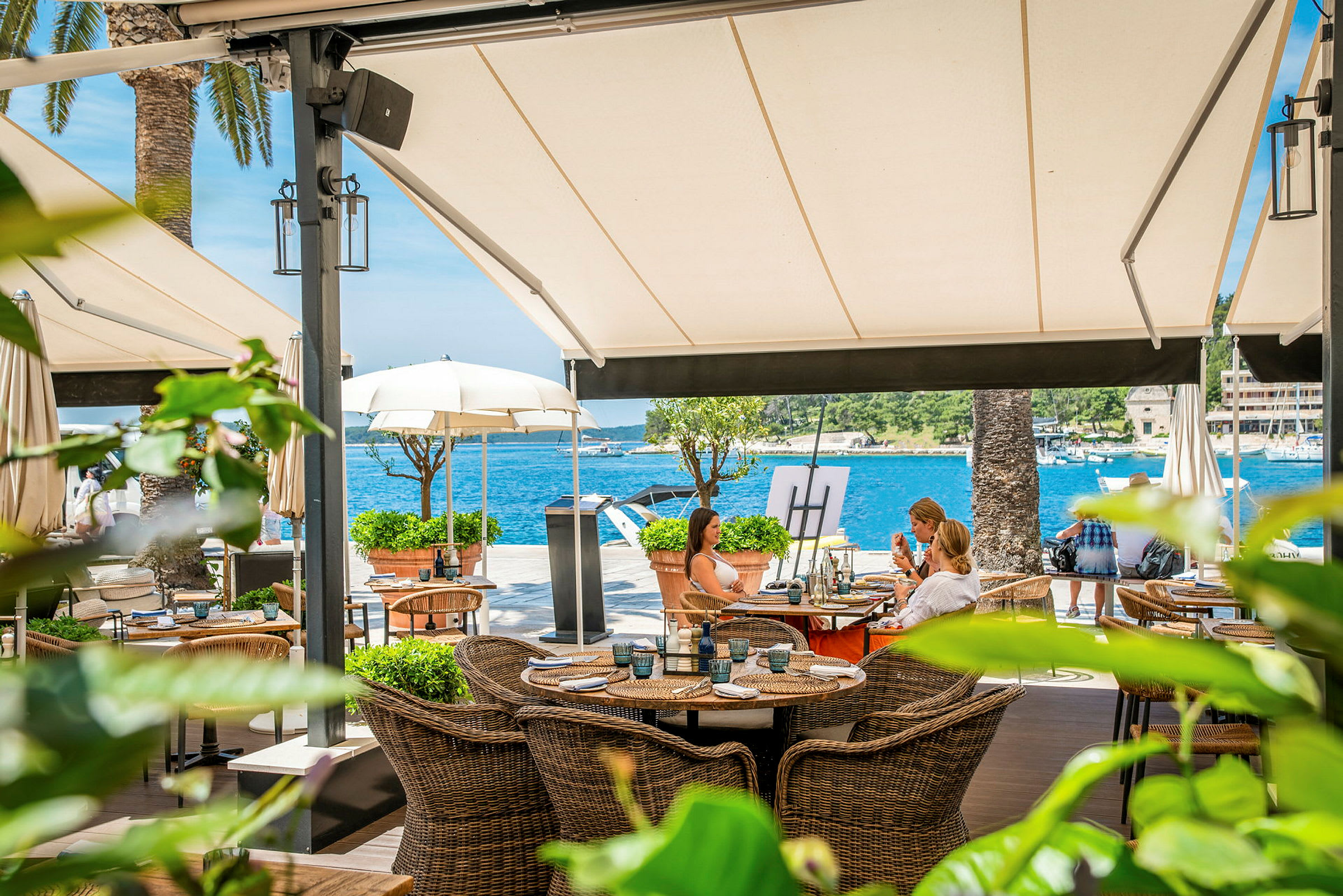 En restaurantterrasse ved vannet med kurvmøbler og utsikt over havet under store markiser. (AI-generert tekst)