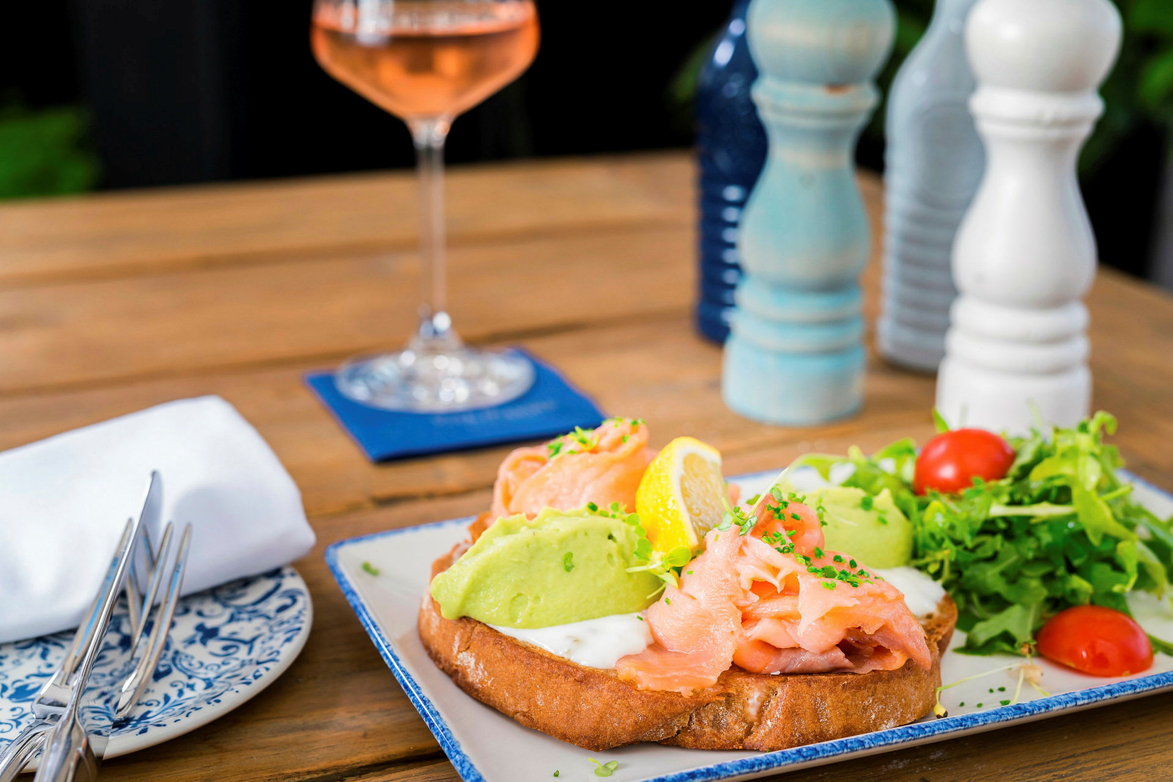 En tallerken med mat: smørbrød med laks, avocado og grønnsaker. I bakgrunnen er det et vinglass og krydderkverner. (AI-generert tekst)