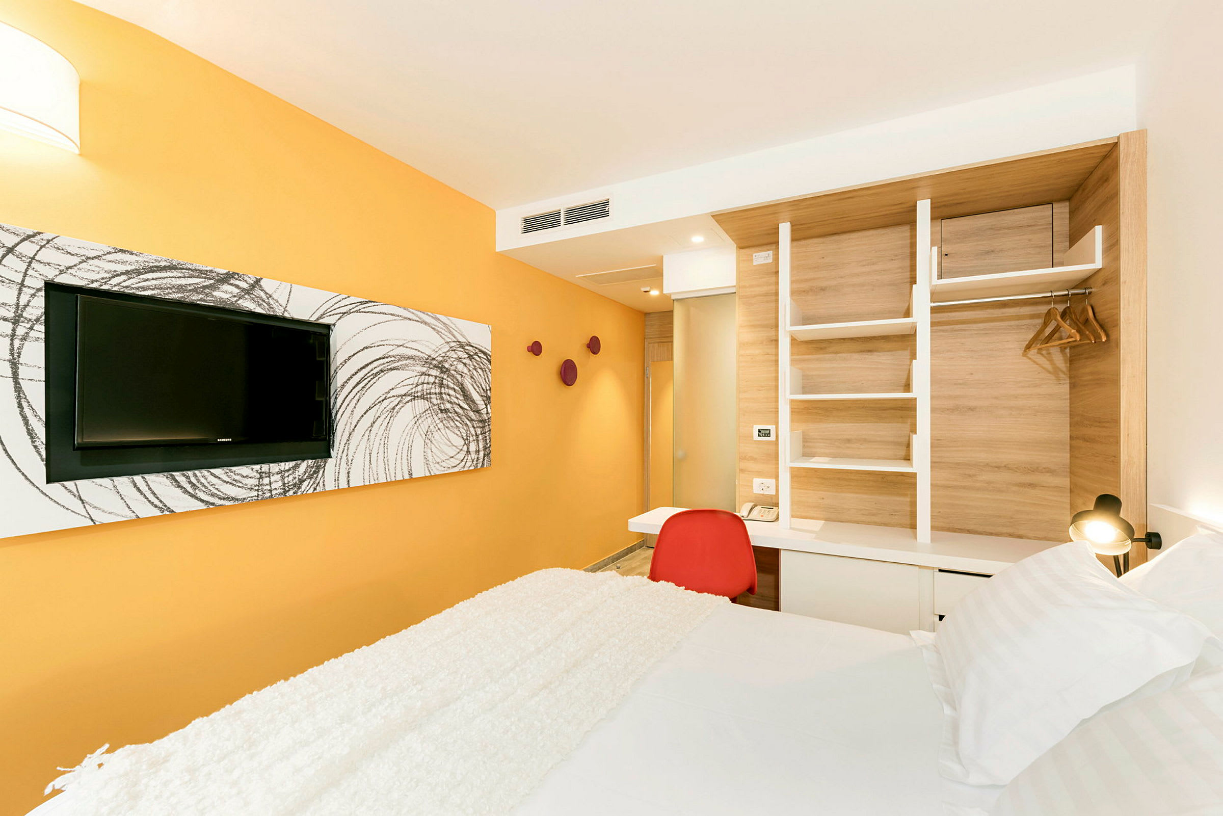 Et hotellrom med en gul vegg, en TV på veggen, en rød stol ved et skrivebord og en seng med hvitt sengetøy. (AI-generert tekst)