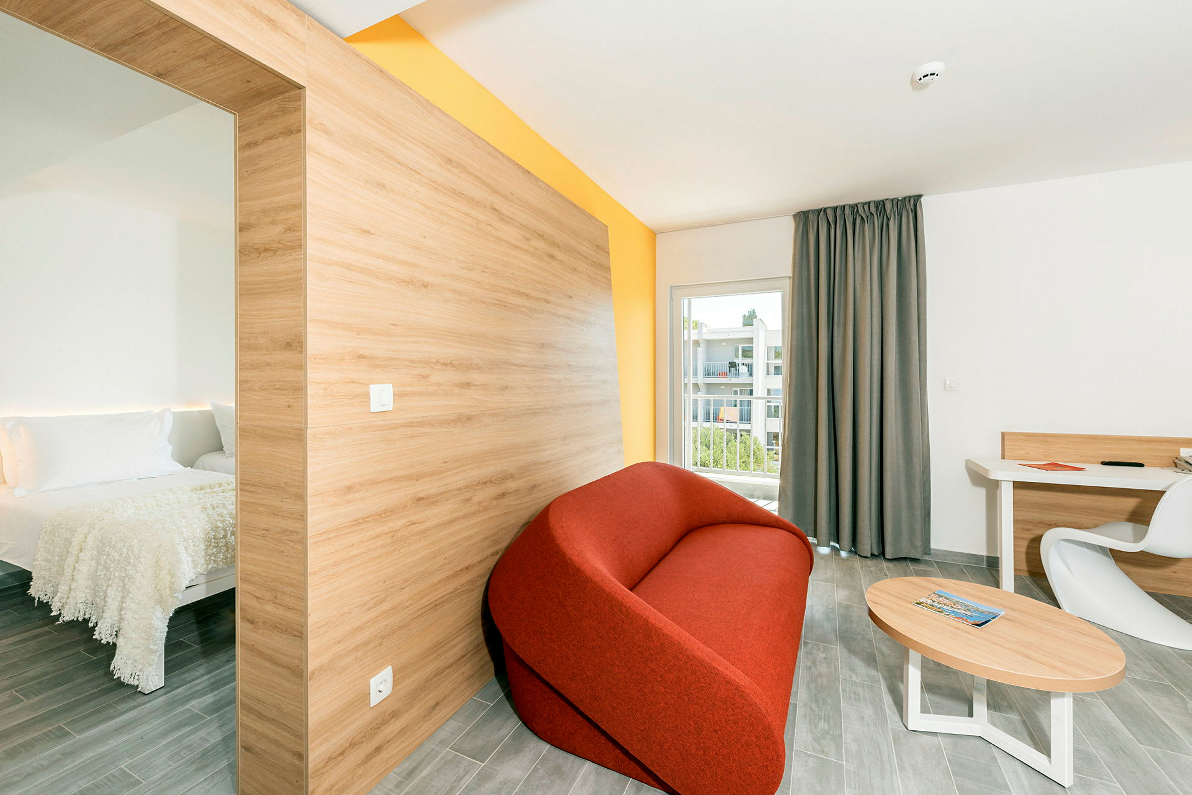 Et moderne hotellrom med et separat soverom til venstre, en rød sofa, et bord og et skrivebord med en stol, samt en balkong med utsikt. (AI-generert tekst)