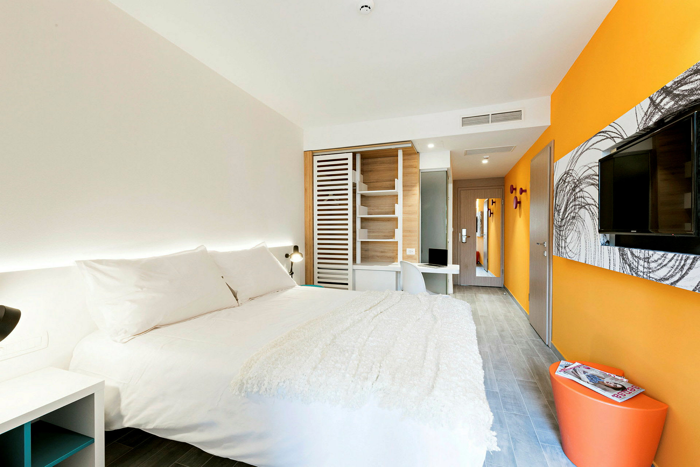 Et moderne hotellrom med en stor seng med hvitt sengetøy, oransje og hvite vegger, en veggmontert TV og et skrivebord. (AI-generert tekst)