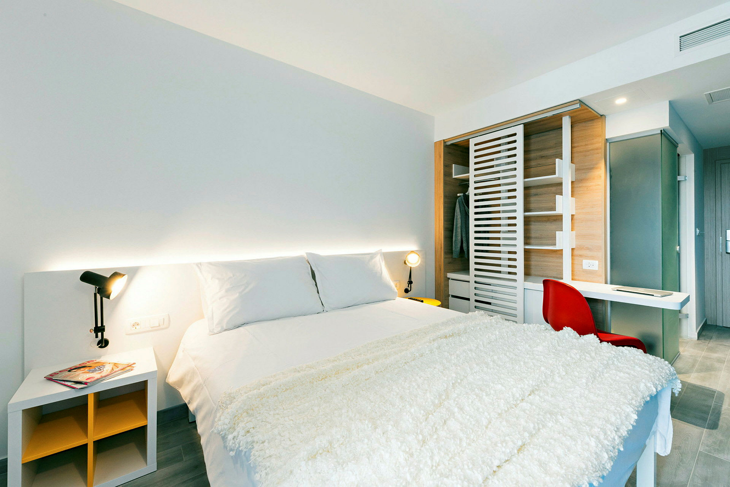 Et moderne soverom med en dobbeltseng, hvitt sengetøy og et pledd. Det er et nattbord med en lampe ved siden av, og et skap er synlig i bakgrunnen. (AI-generert tekst)