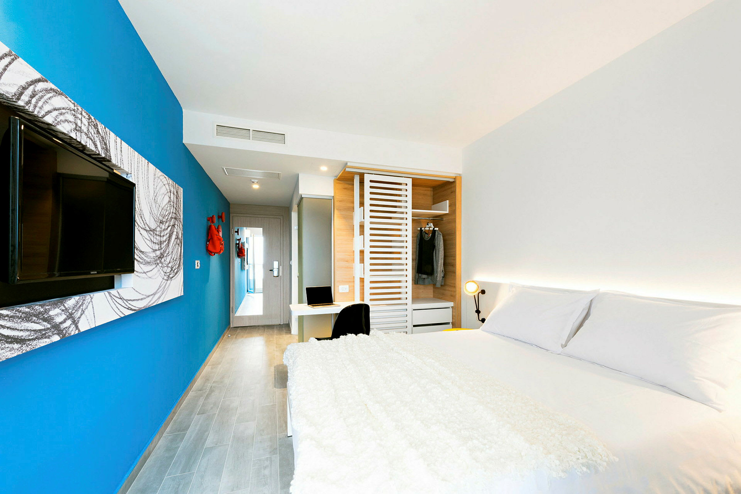 Et moderne hotellrom med en stor seng, blå vegg, TV, garderobe og et skrivebord. (AI-generert tekst)
