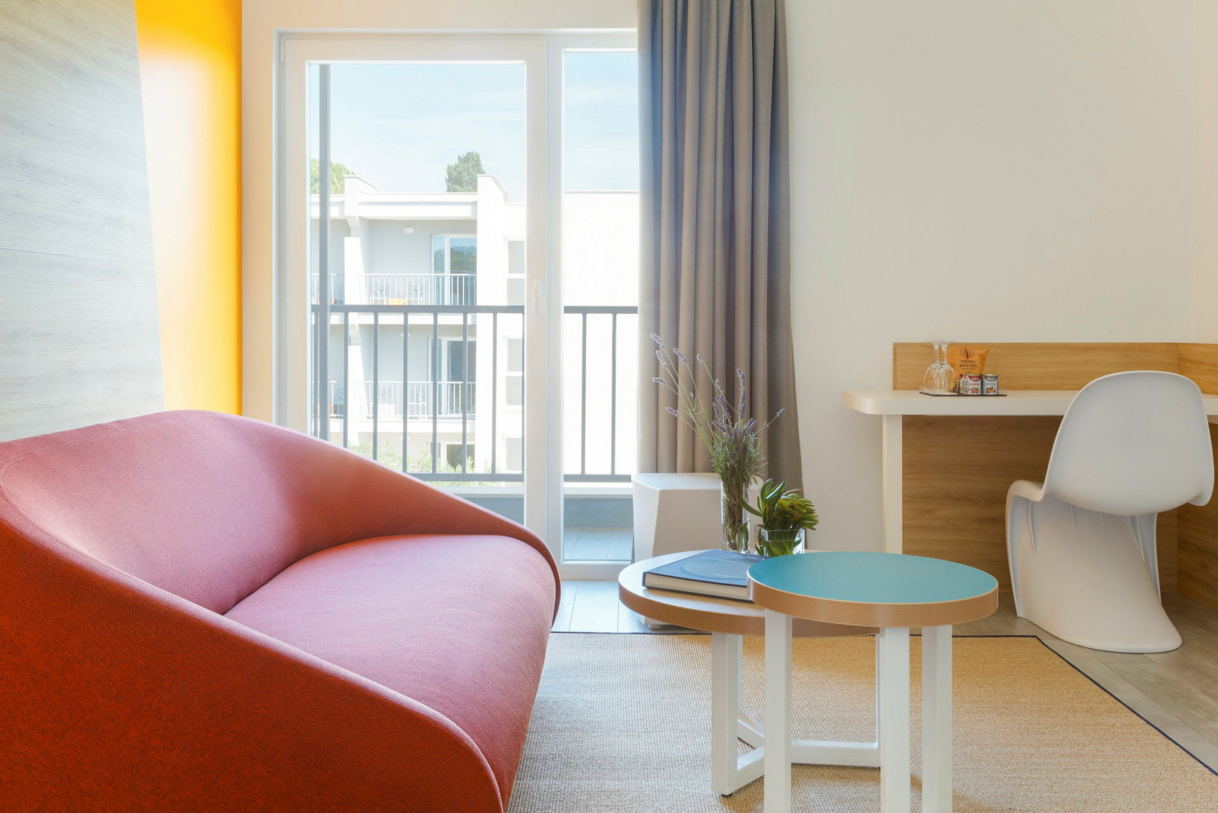 En moderne innredet hotellromshjørne med en rød sofa, et lite bord med dekorasjoner og en skrivebordskrok ved et stort vindu. (AI-generert tekst)