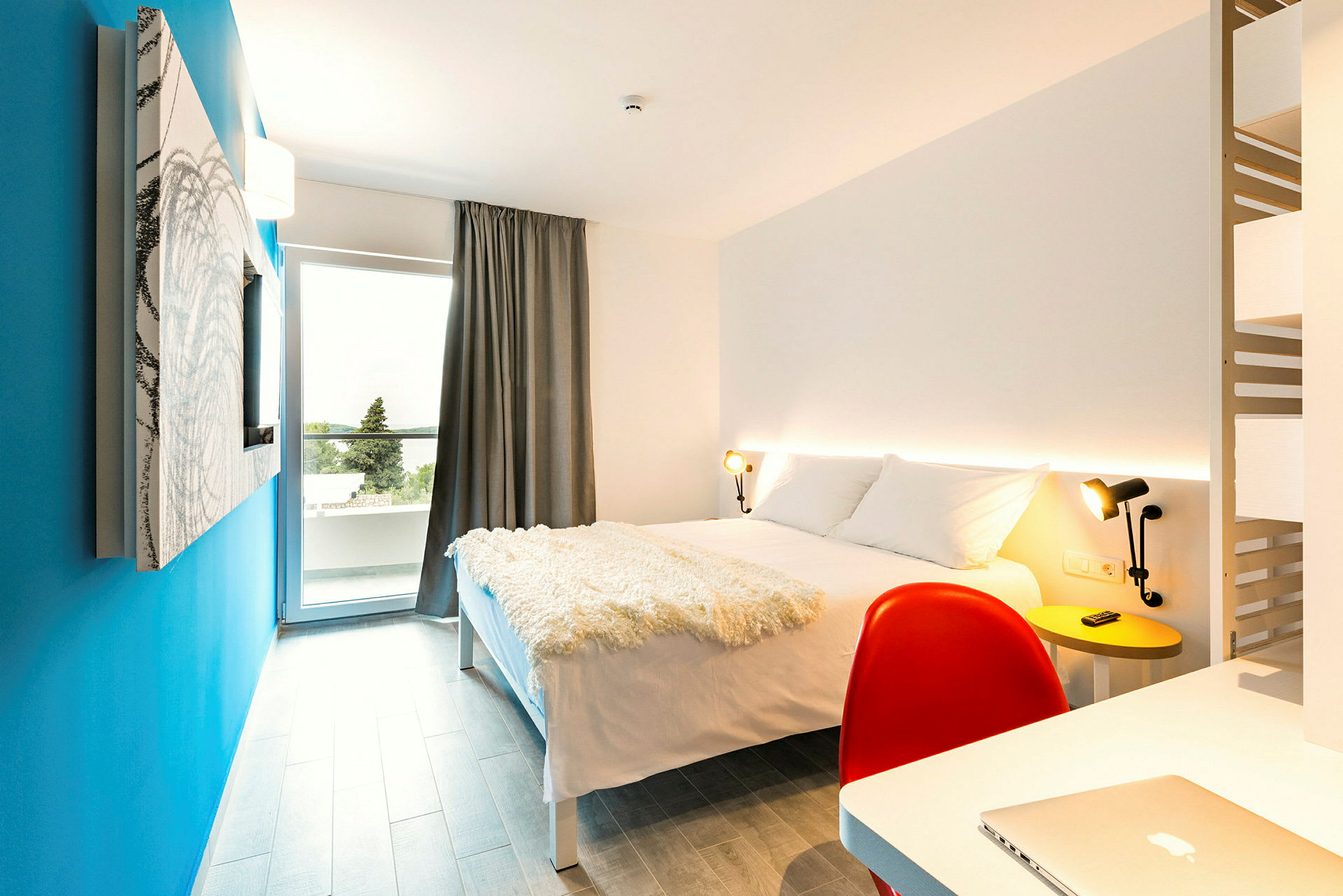 Moderne utstyrt hotellrom med en dobbeltseng, skrivebord og en blå vegg. (AI-generert tekst)