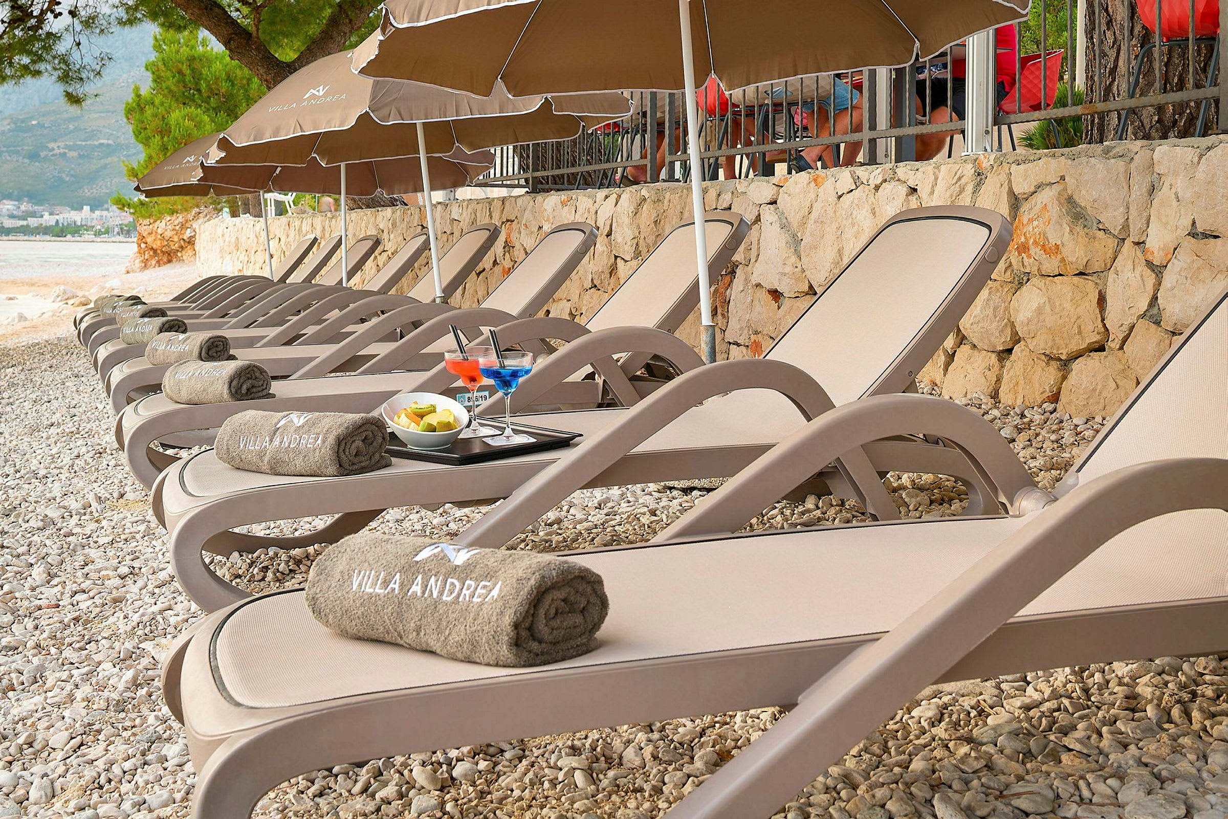 Rad med solsenger på en steinstrand, med håndklær merket 'Villa Andrea', drinker og frukt på et brett. (AI-generert tekst)