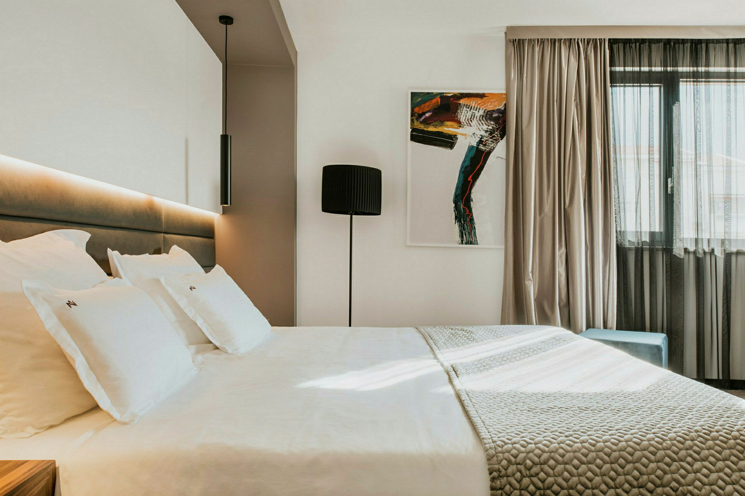 Moderne hotellrom med en stor seng, kunstverk på veggen og et vindu med lyse gardiner. (AI-generert tekst)