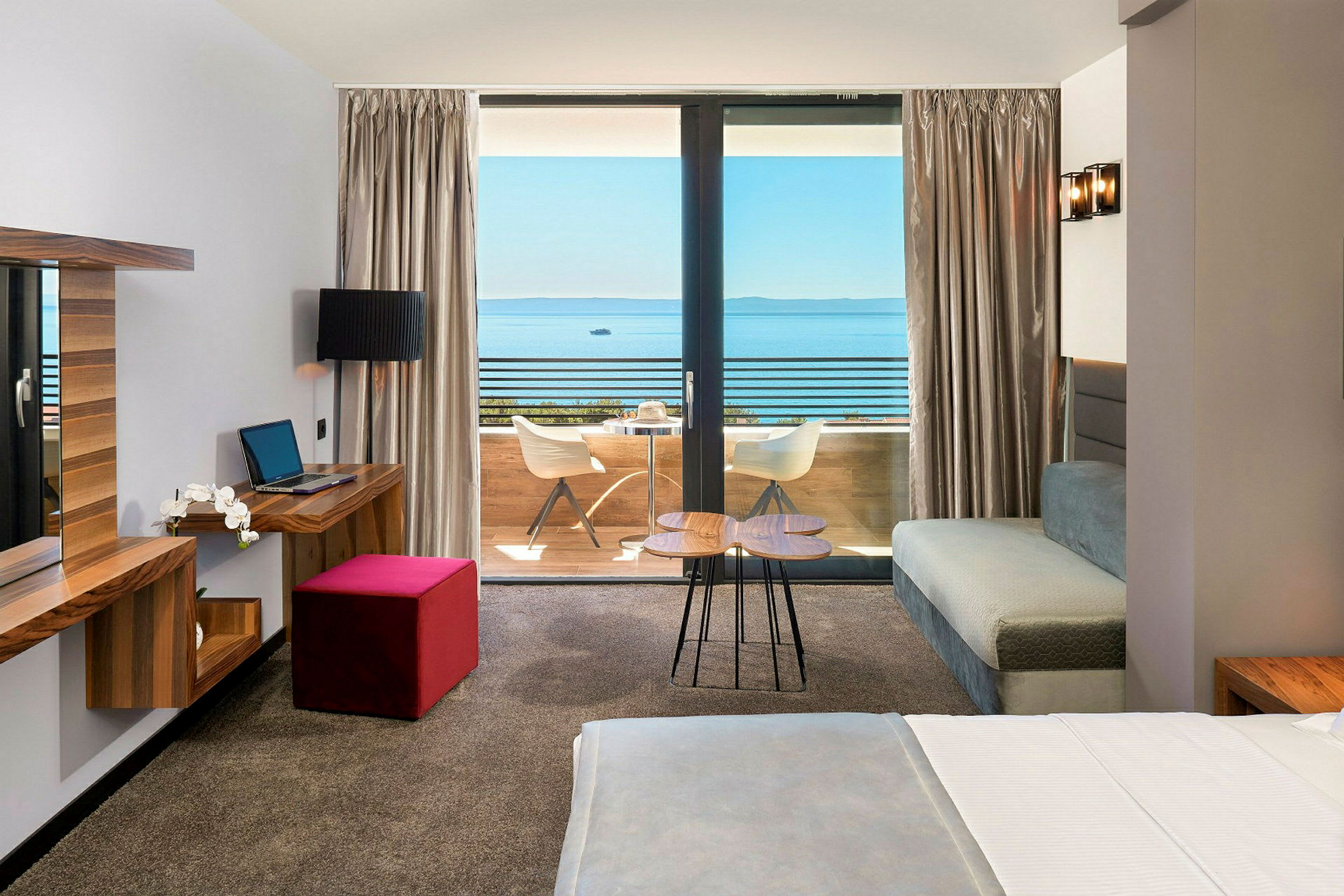 Hotellrom med seng, skrivebord og utsikt mot en balkong og havet. (AI-generert tekst)