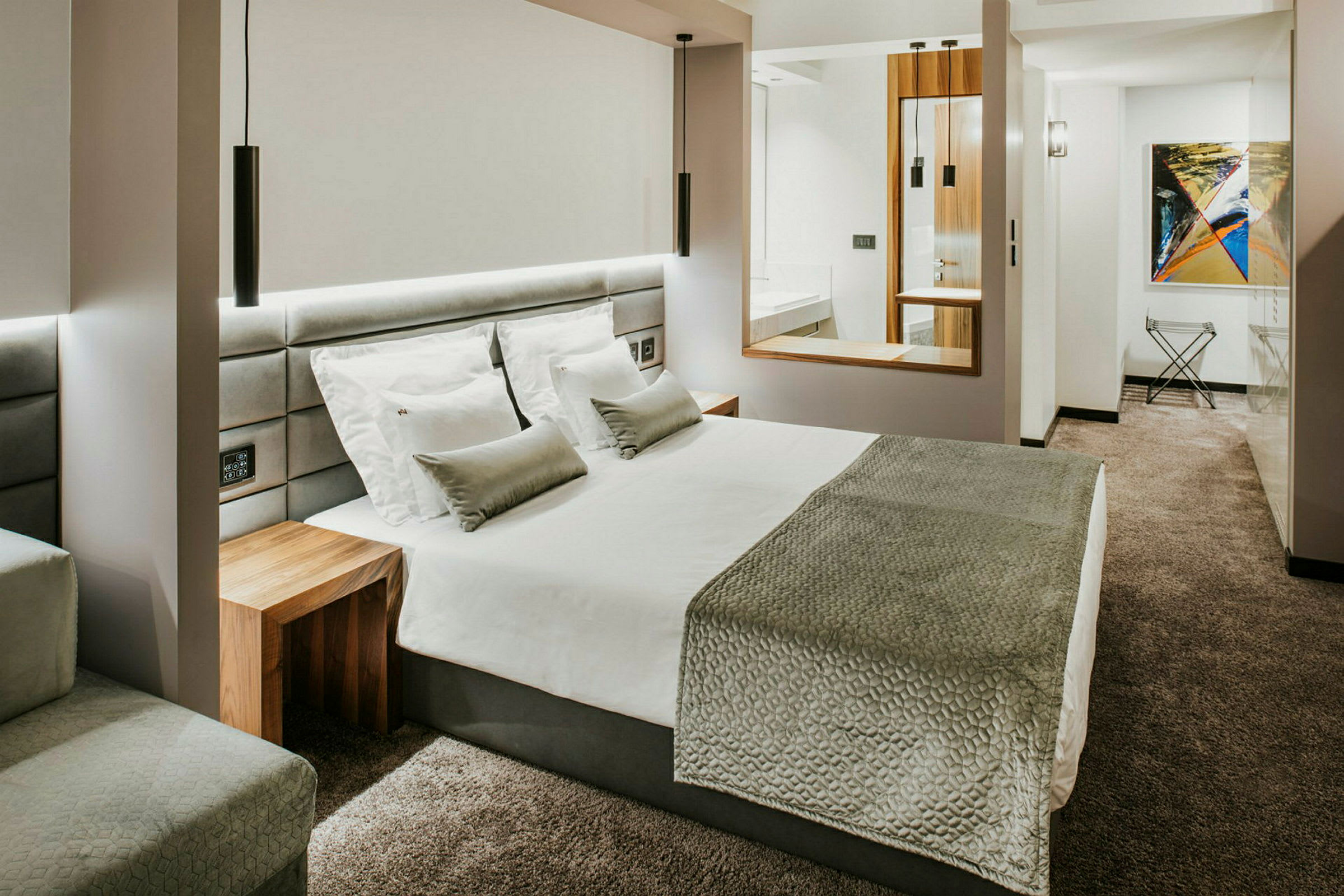 Et moderne hotellrom med en stor seng, hvite sengeklær, grått sengeteppe og puter, og en grå vegg-til-vegg teppe. (AI-generert tekst)