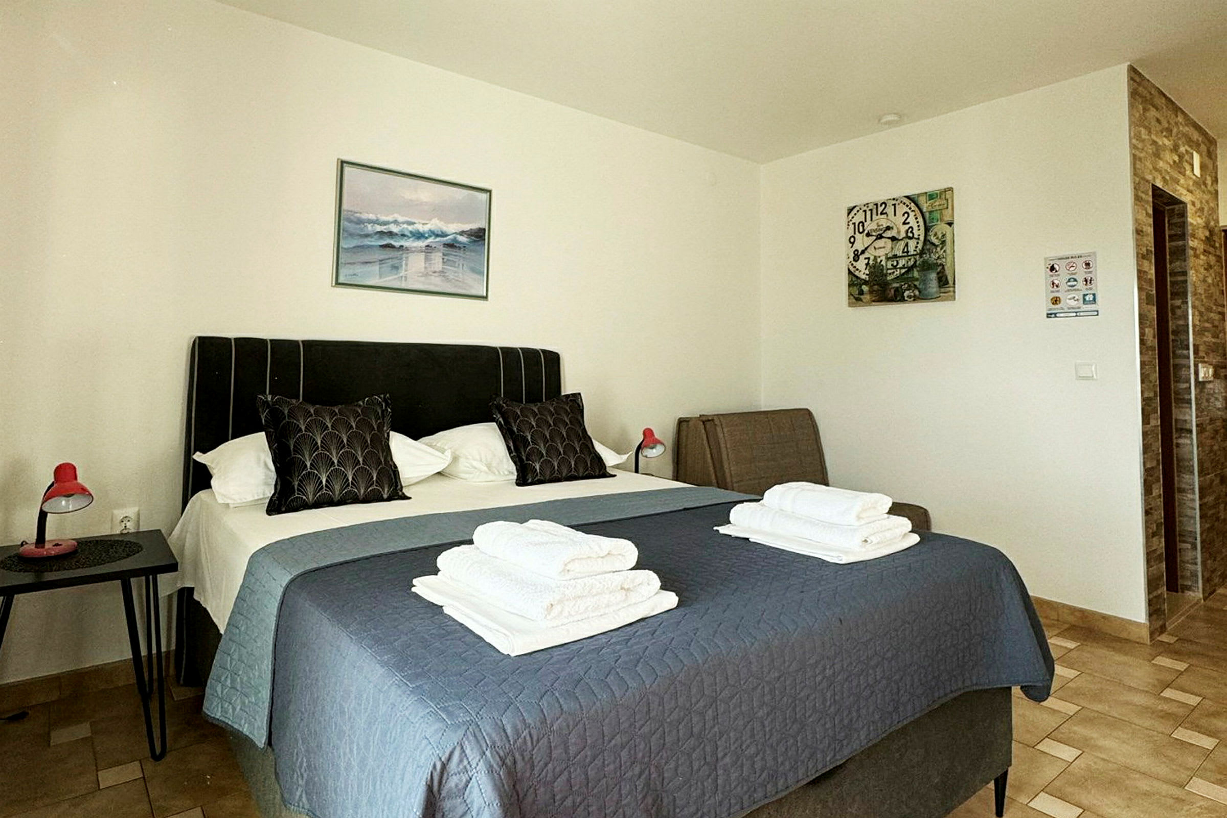 Et hotellrom med en seng dekket med et blått sengeteppe, to puter og sammenbrettede håndklær oppå. Malerier henger på veggene, og et nattbord med en rød lampe står ved siden av sengen. (AI-generert tekst)