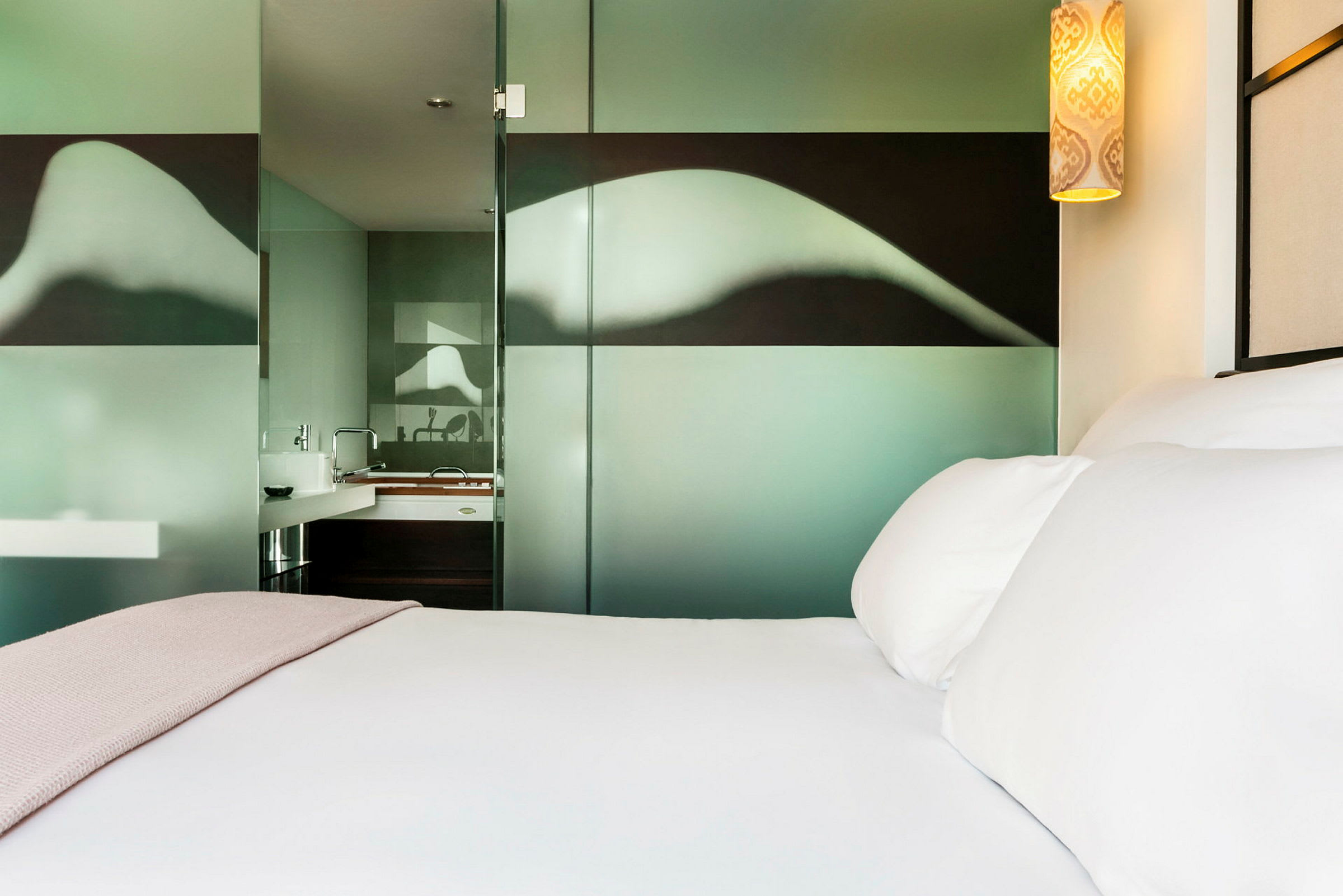 Et moderne hotellrom viser en seng med hvitt sengetøy og et rosa teppe, samt en glassvegg med mønster som skiller badet. (AI-generert tekst)