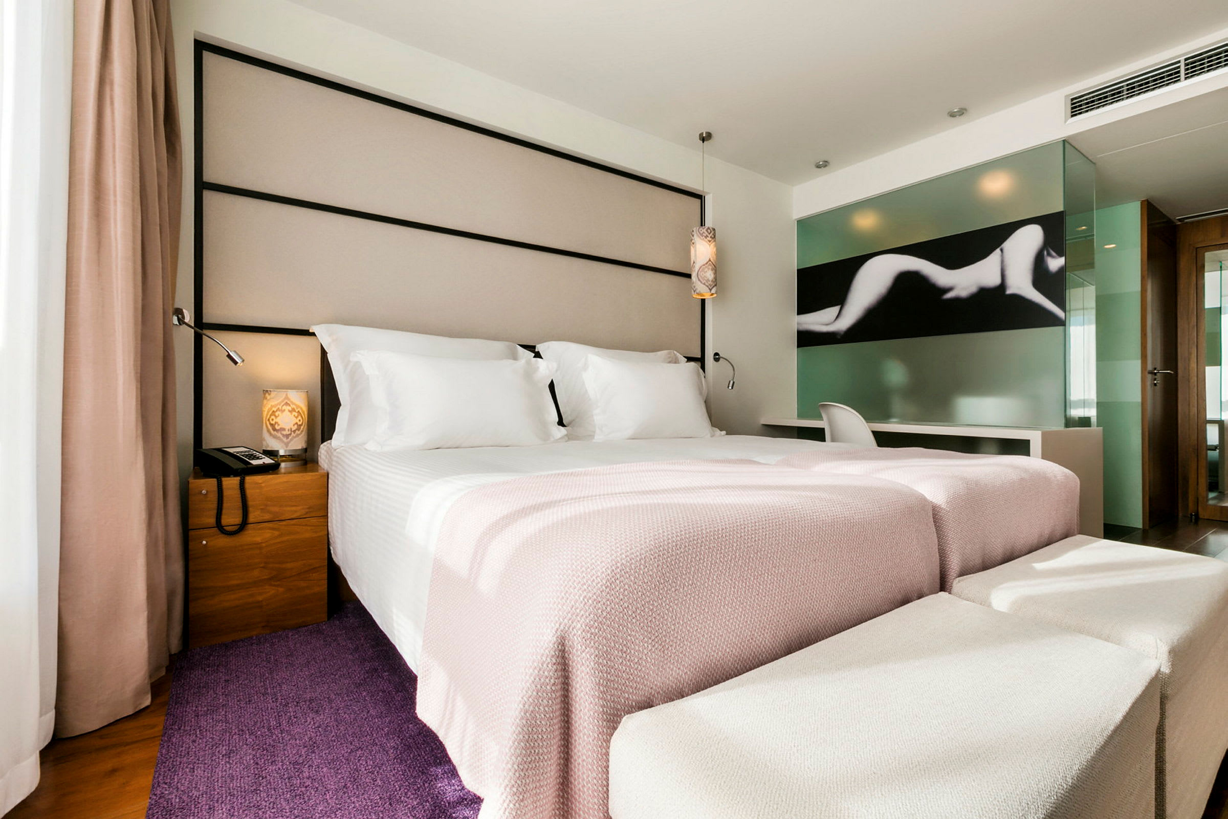 Et moderne hotellrom med en bred seng oppredd med hvitt og lysrosa sengetøy. Det er en stol og et skrivebord ved en glassvegg med kunstmotiv. (AI-generert tekst)