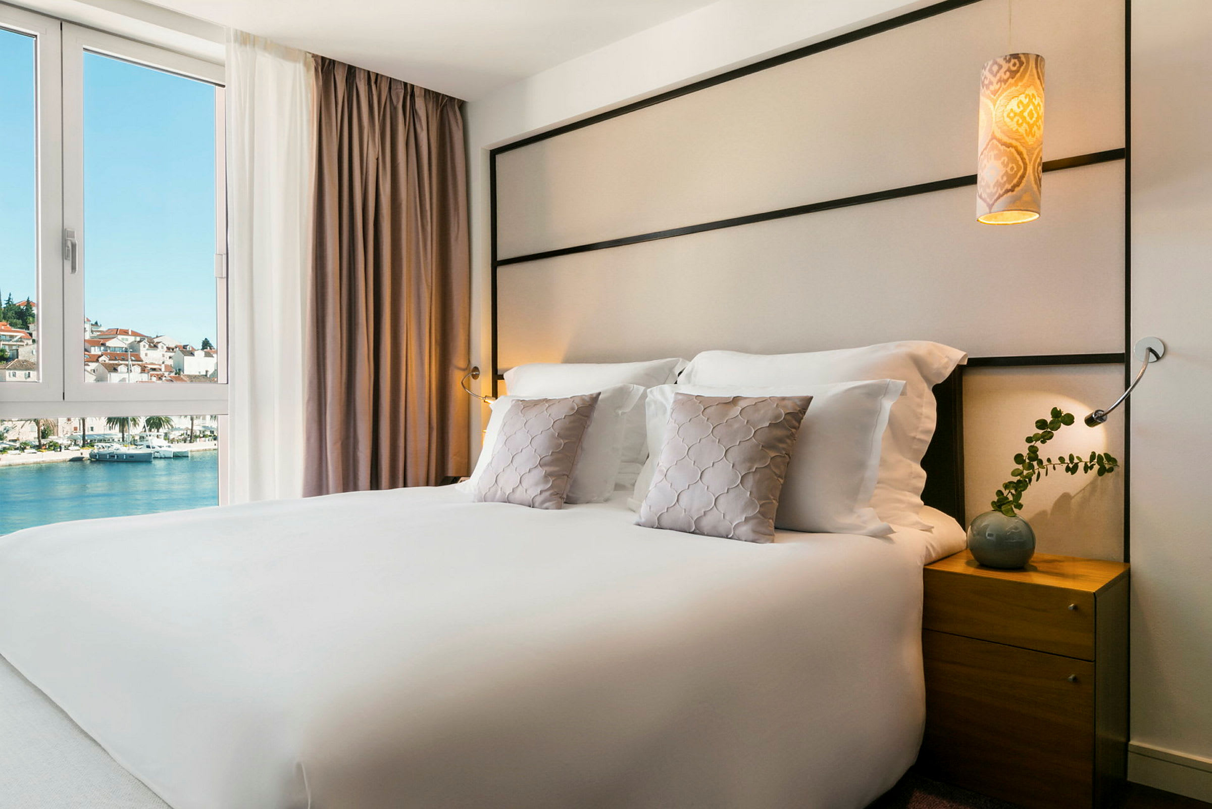 Hotellrom med en dobbeltseng, pyntputer og et nattbord. Utsikt over vann og bygninger gjennom vinduet. (AI-generert tekst)