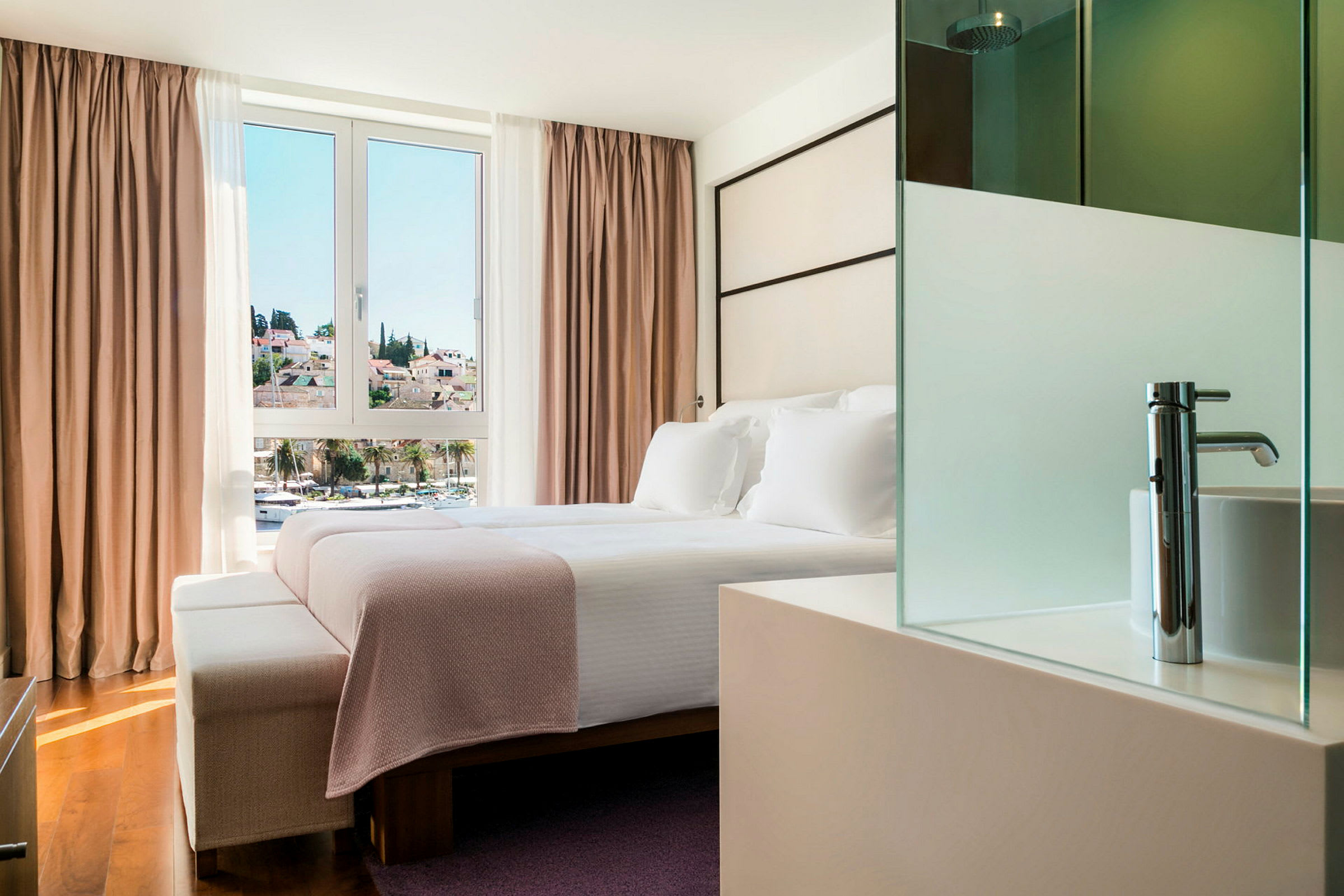 Et hotellrom med en dobbeltseng plassert foran et stort vindu med utsikt over en by. Sengen er redd opp med hvitt sengetøy og et rosa pledd. (AI-generert tekst)