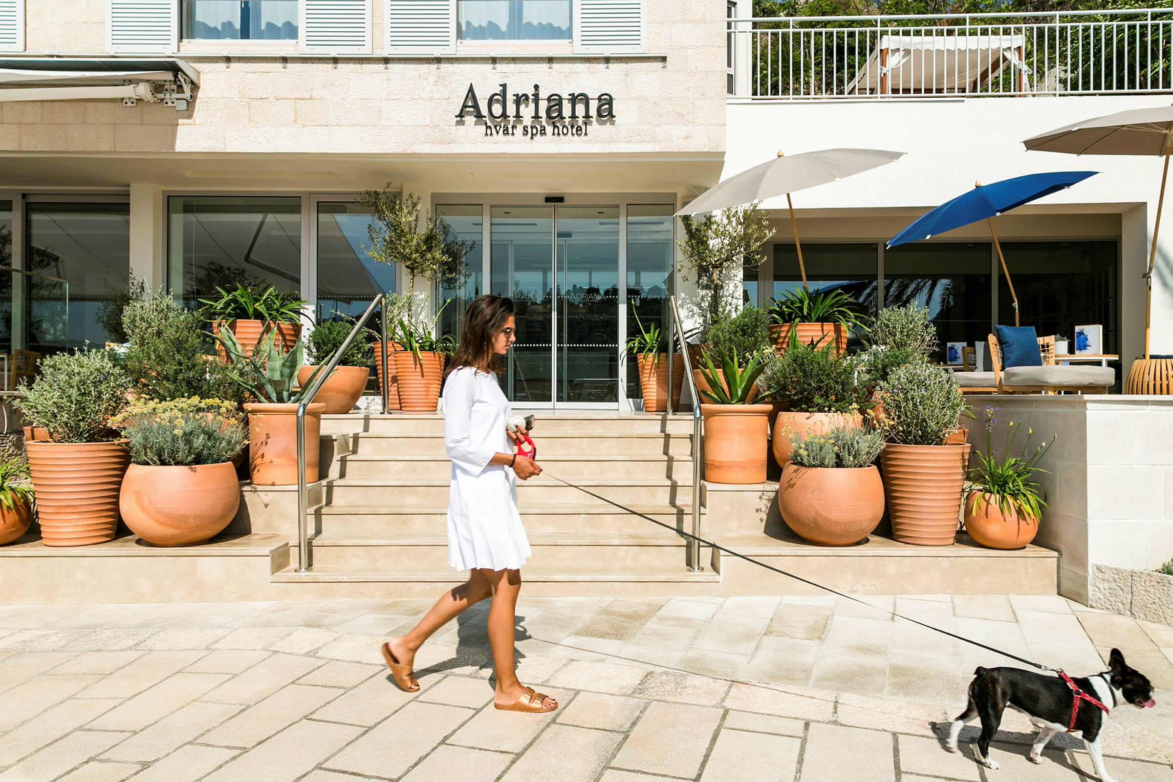 En kvinne går med en hund i bånd foran inngangen til Adriana Hvar Spa Hotel. (AI-generert tekst)
