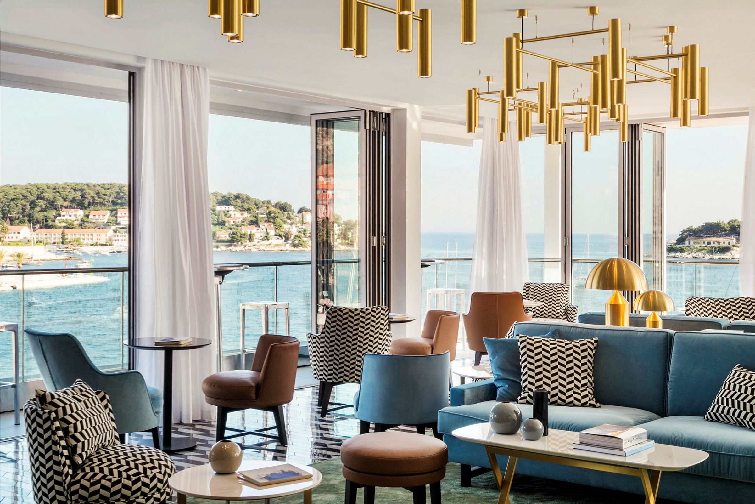 En moderne lounge med sittemøbler og dekorative lysekroner, omgitt av store vinduer som gir utsikt over en kystby og havet. (AI-generert tekst)