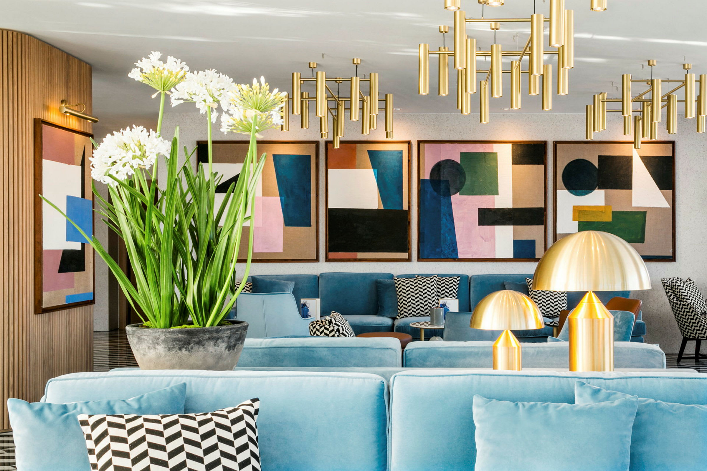 Et moderne innredet rom med lyseblå sofaer, dekorative puter med grafiske mønstre og store metalliske lamper. Veggene er prydet med abstrakt kunst, og en grønn plante med hvite blomster står på et bord. (AI-generert tekst)