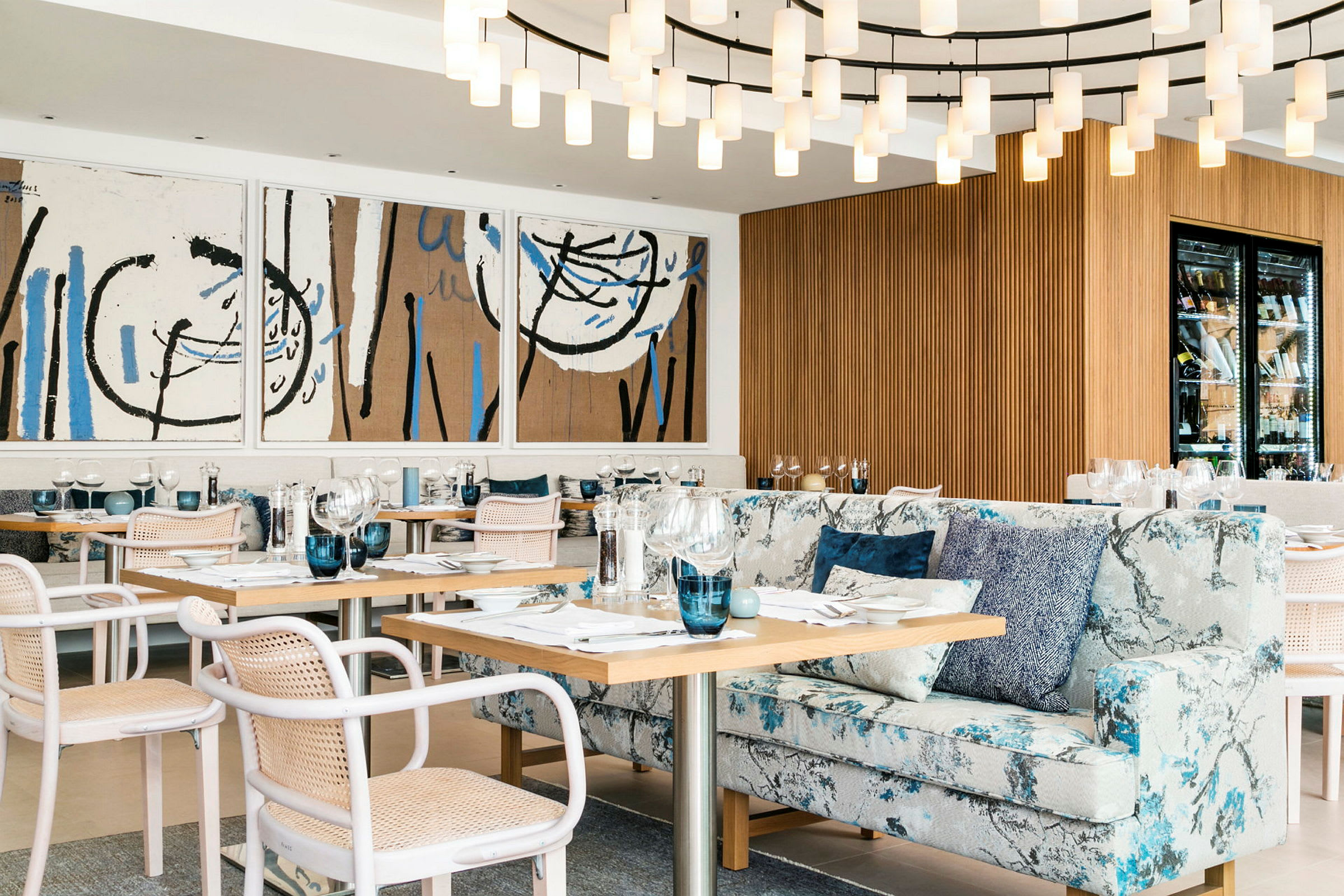 Et elegant innredet restaurantområde med moderne kunstverk på veggen, en stor lysekrone, komfortable stoler og sofaer samt dekkede bord. (AI-generert tekst)