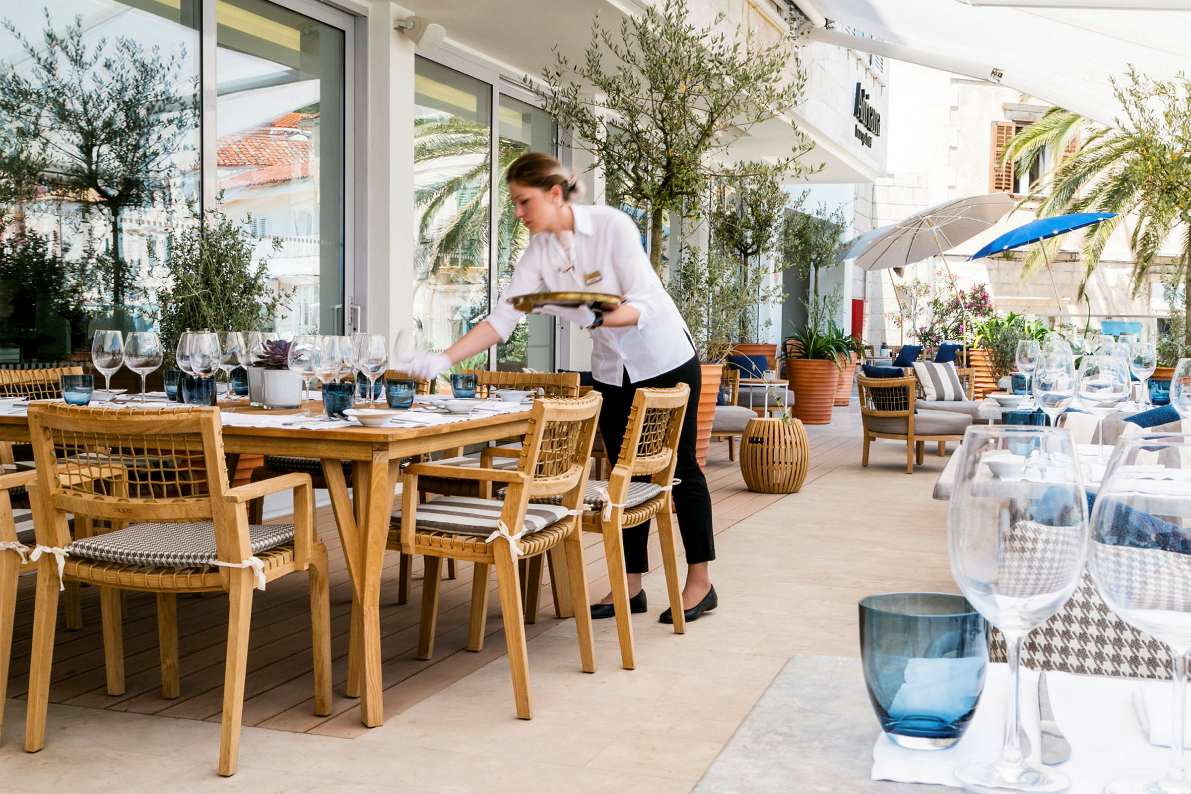 En utendørs restaurant med trebord og stoler samt oppdekning med glass og servise. Vakker omgivelser med planter og parasoller. (AI-generert tekst)