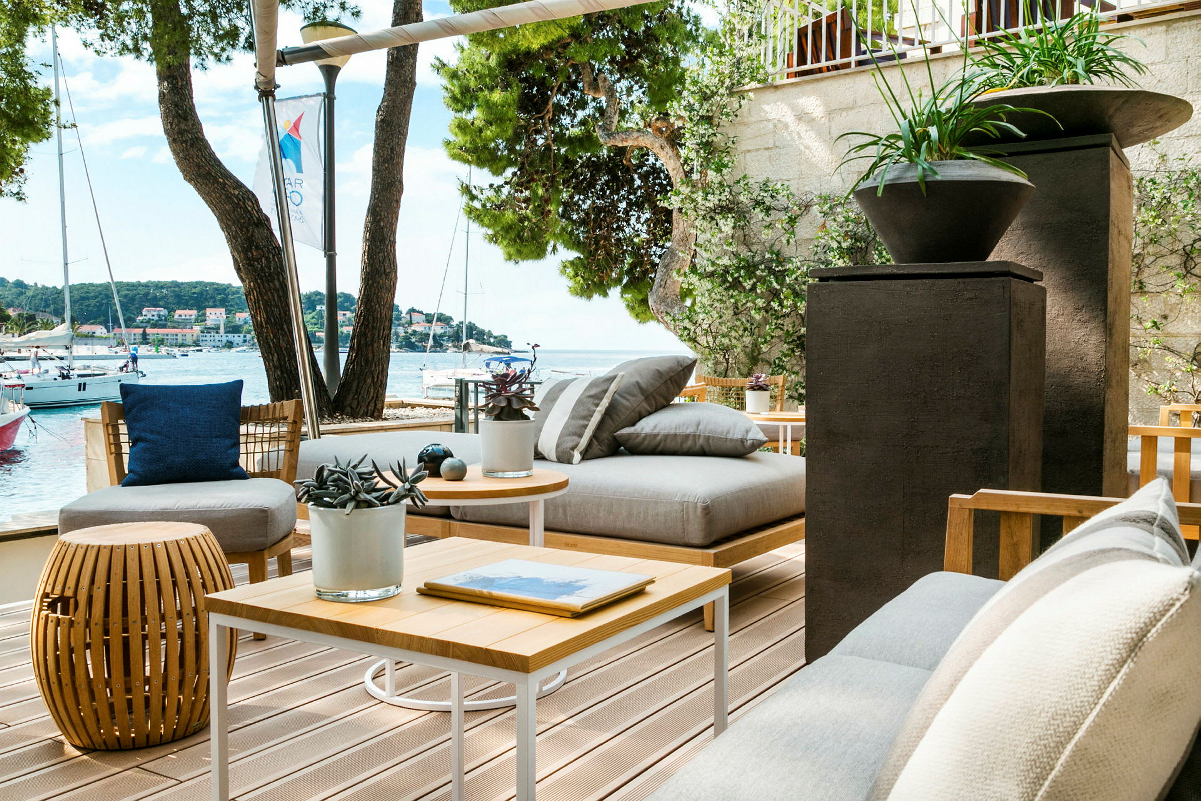 Et utendørsområde med moderne sittemøbler, planter i potter og utsikt over havet med seilbåter. (AI-generert tekst)