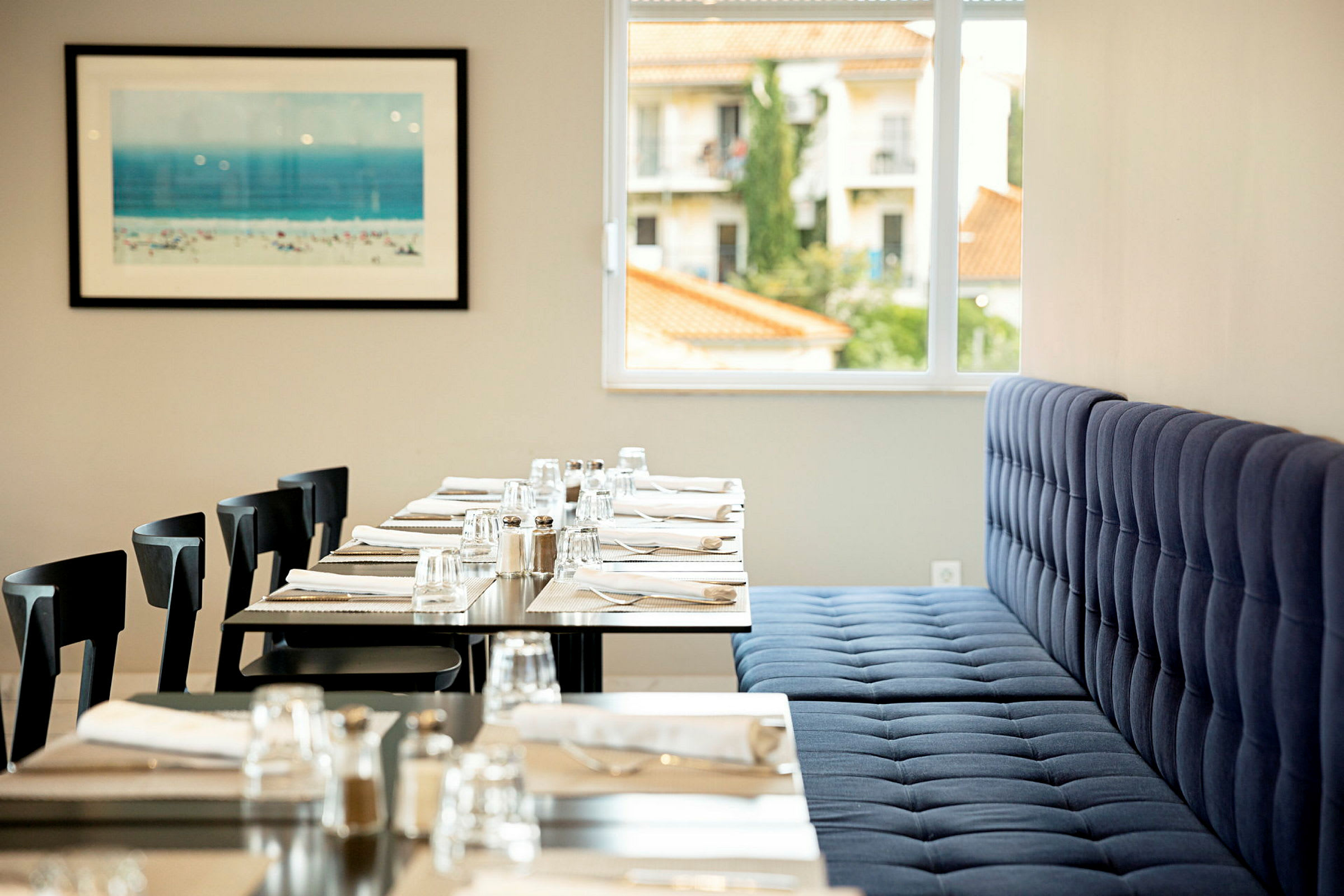 Et restaurantmiljø med dekkede bord med glass, bestikk og servietter. En blå polstret sofa og vindu som viser bygninger utenfor. Et maleri med strandmotiv på veggen. (AI-generert tekst)