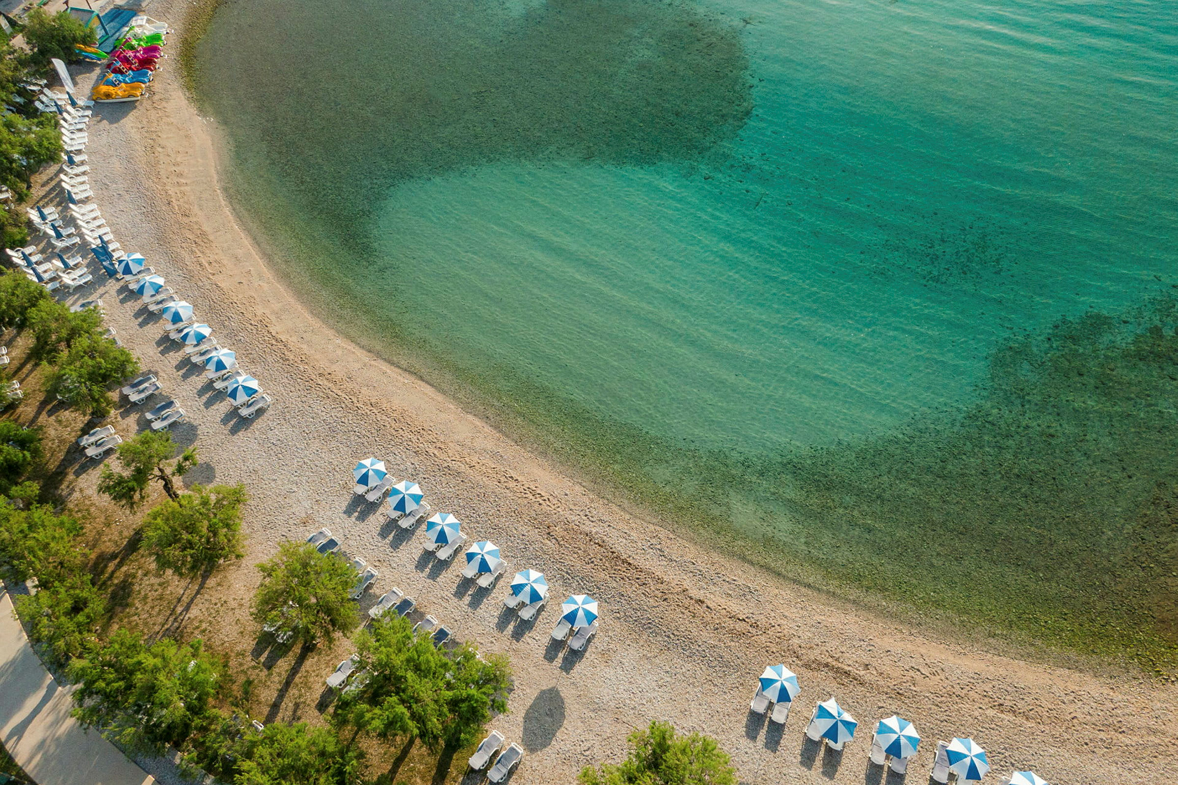 Flyutsikt over strand med parasoller og solsenger ved klart vann. (AI-generert tekst)