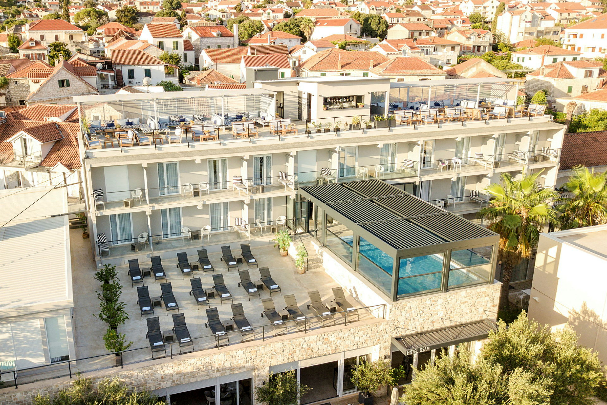 Utsikt over hotell med basseng, solsenger og takterrasse i en bymiljø. (AI-generert tekst)