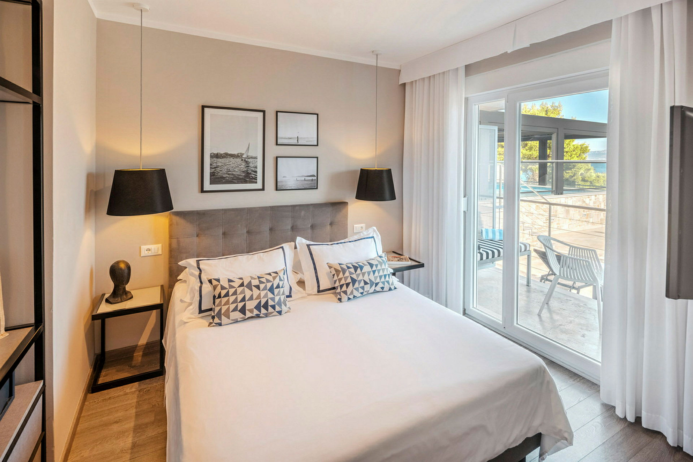 Moderne hotellrom med dobbeltseng, dekorert med puter og kunst på veggen. Glassdører fører ut til en møblert terrasse. (AI-generert tekst)
