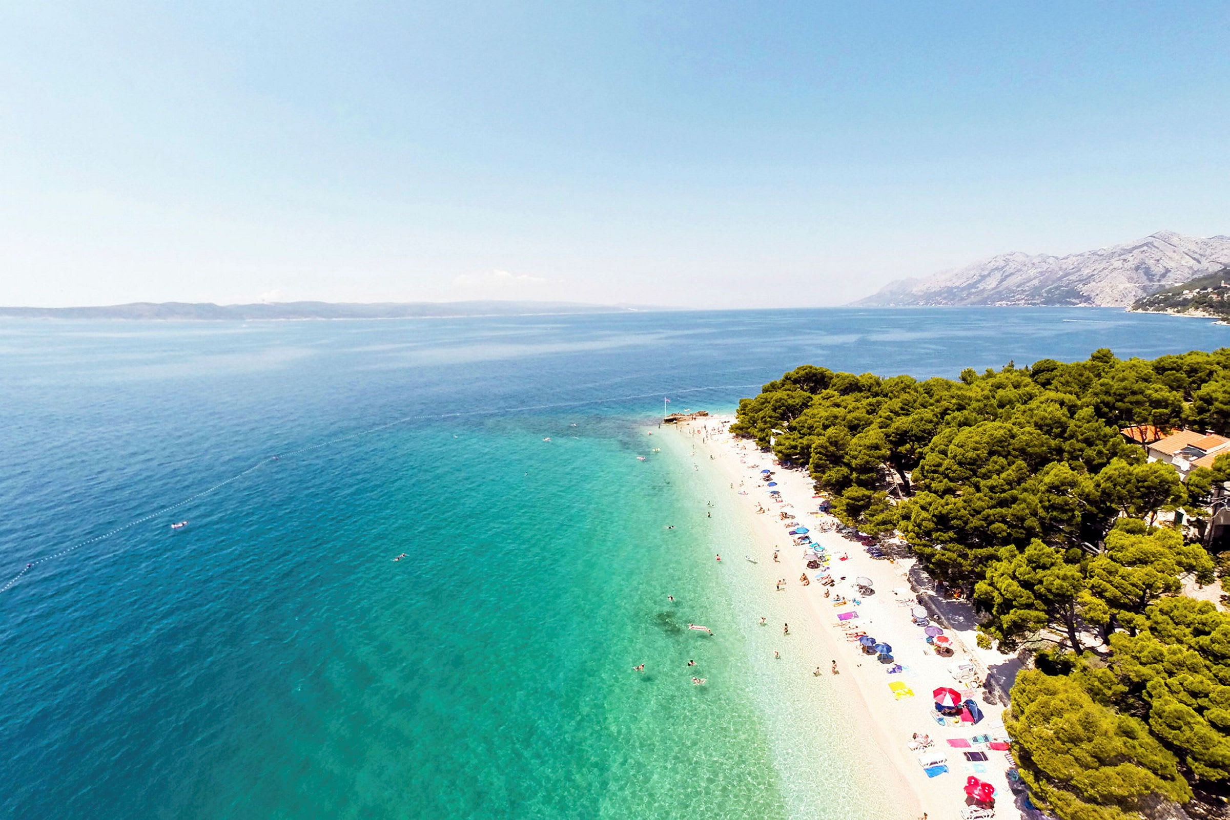 Strand med turkist vann, parasoller og badegjester, omgitt av grønne trær. (AI-generert tekst)