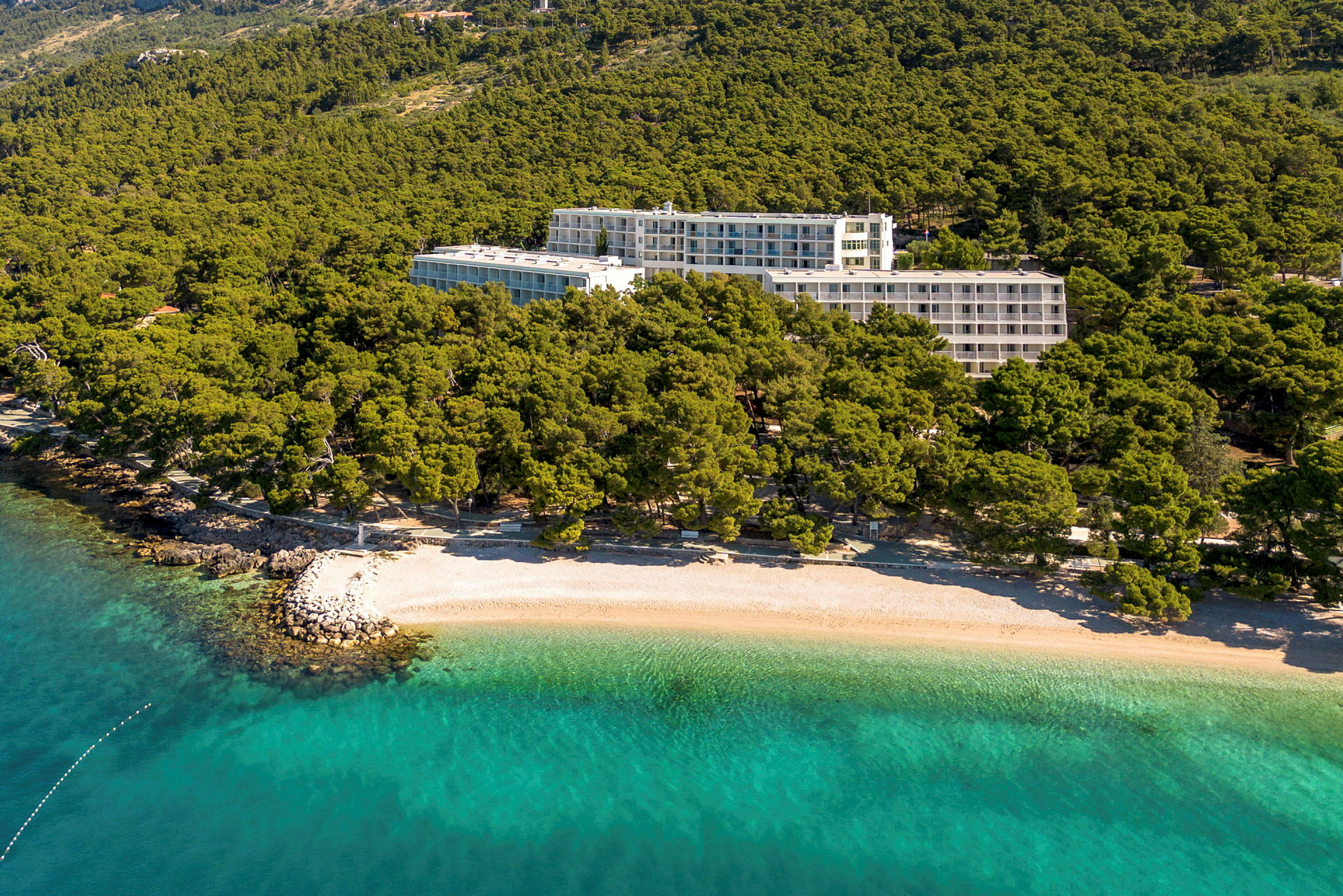 Hotellbygninger omgitt av grøntområder nær en strand med klart turkist vann. (AI-generert tekst)