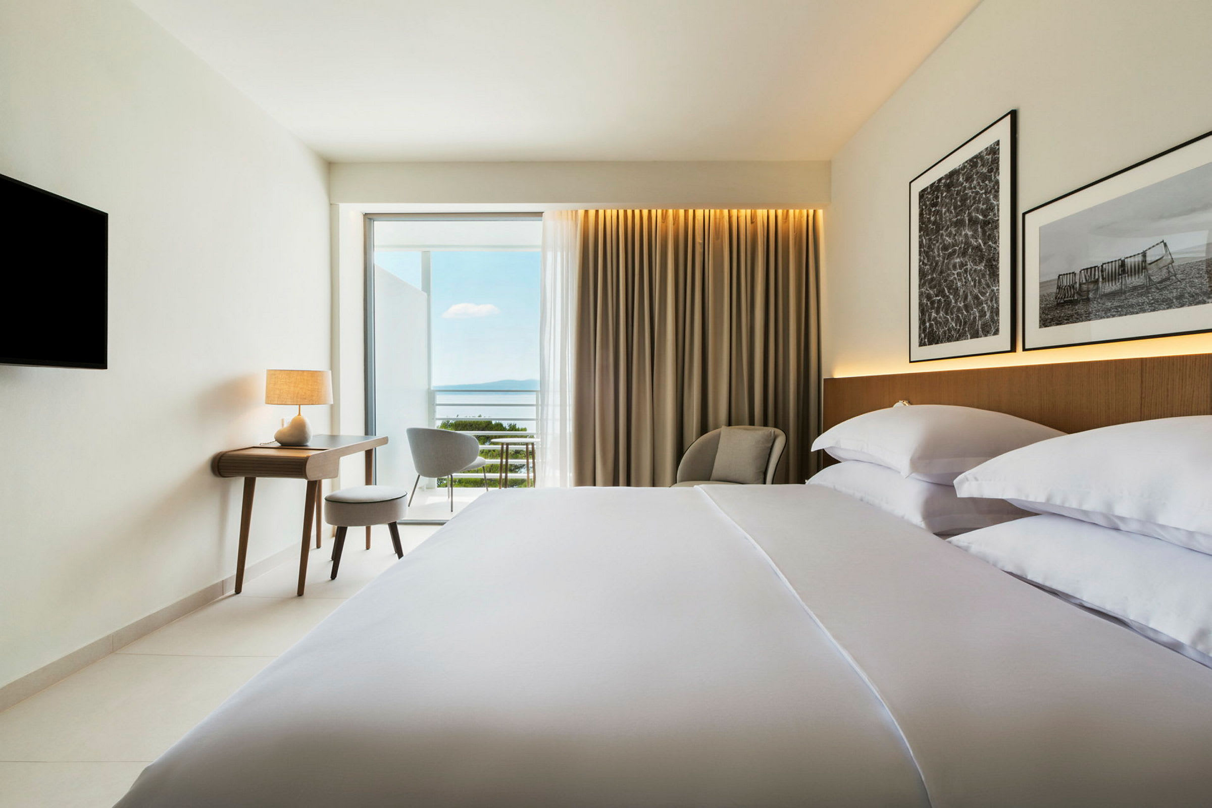 Hotellrom med oppredd seng, skrivebord, stol og utsikt mot havet. (AI-generert tekst)